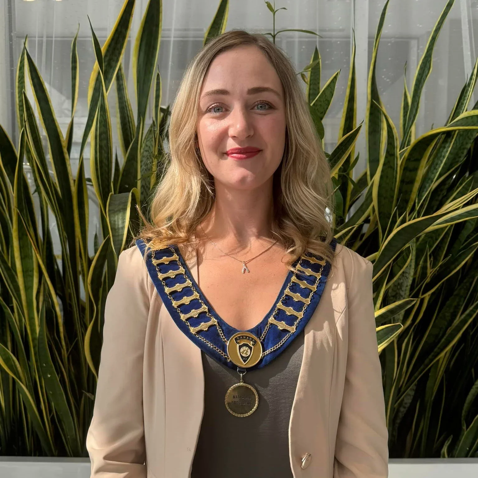 A young woman with blonde hair and blue eyes standing in front of tall green plants, wearing a beige blazer and a sash with medals and chains, smiling at the camera.