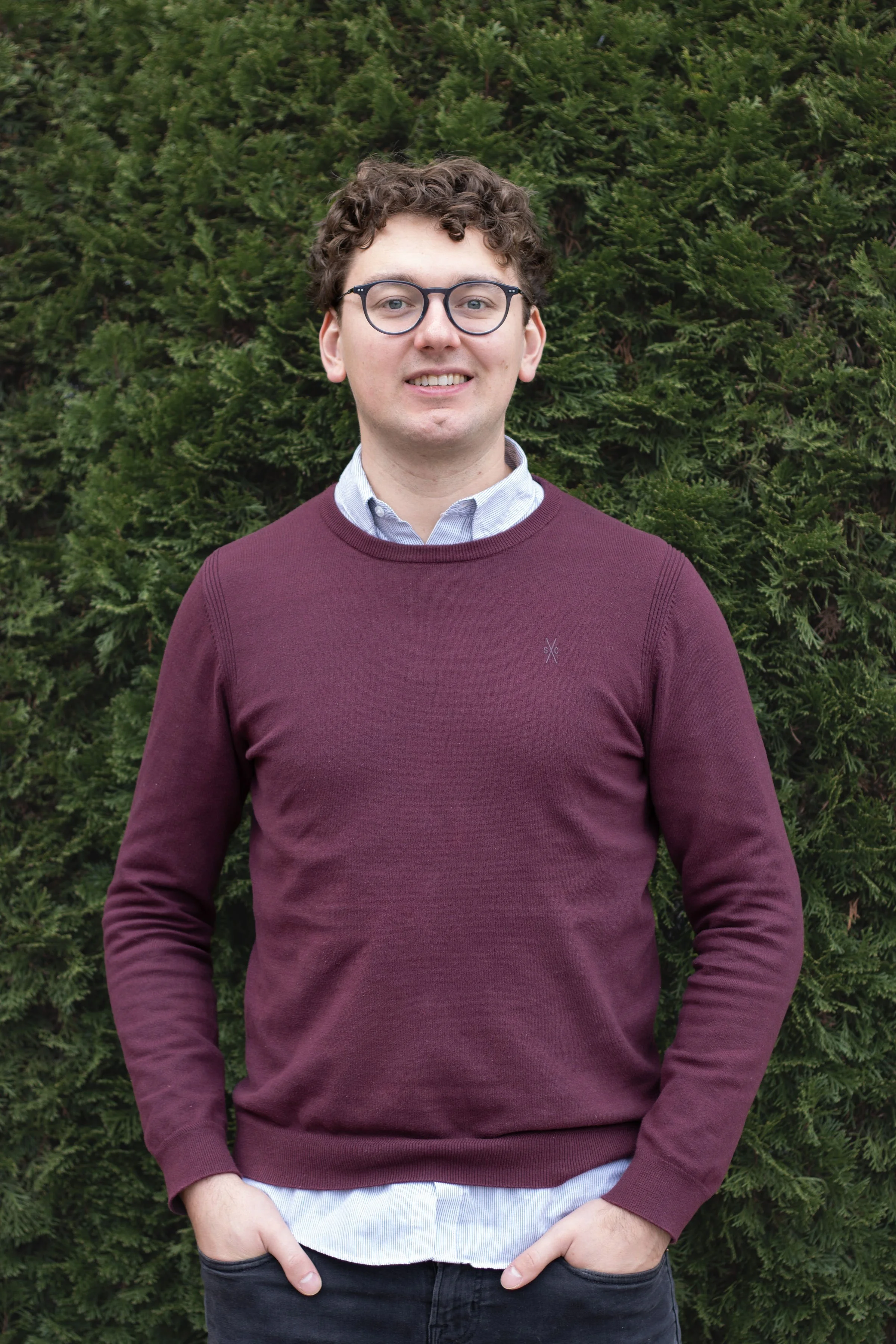 A young man with glasses and curly brown hair standing outdoors in front of a green bush, wearing a maroon sweater over a light-colored collared shirt and black pants.