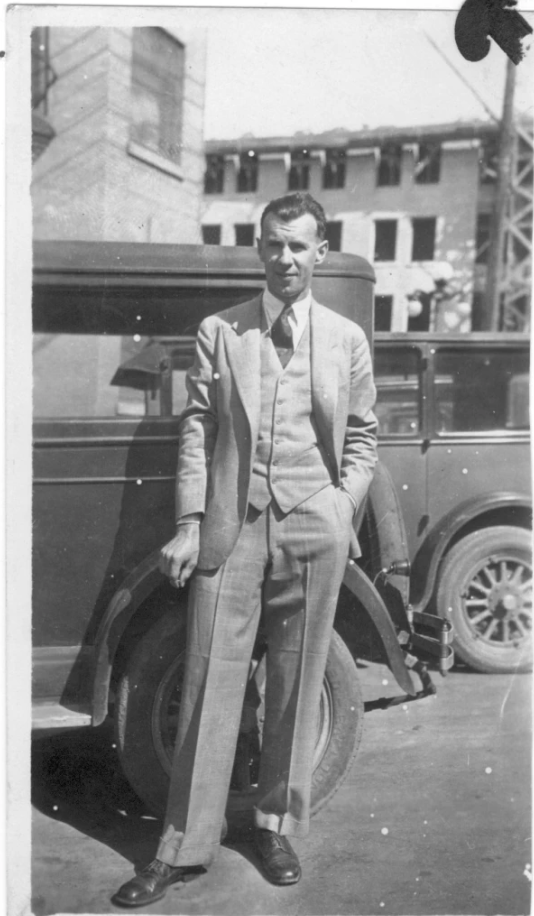 A man in a suit standing next to a vintage car on a city street with buildings in the background.
