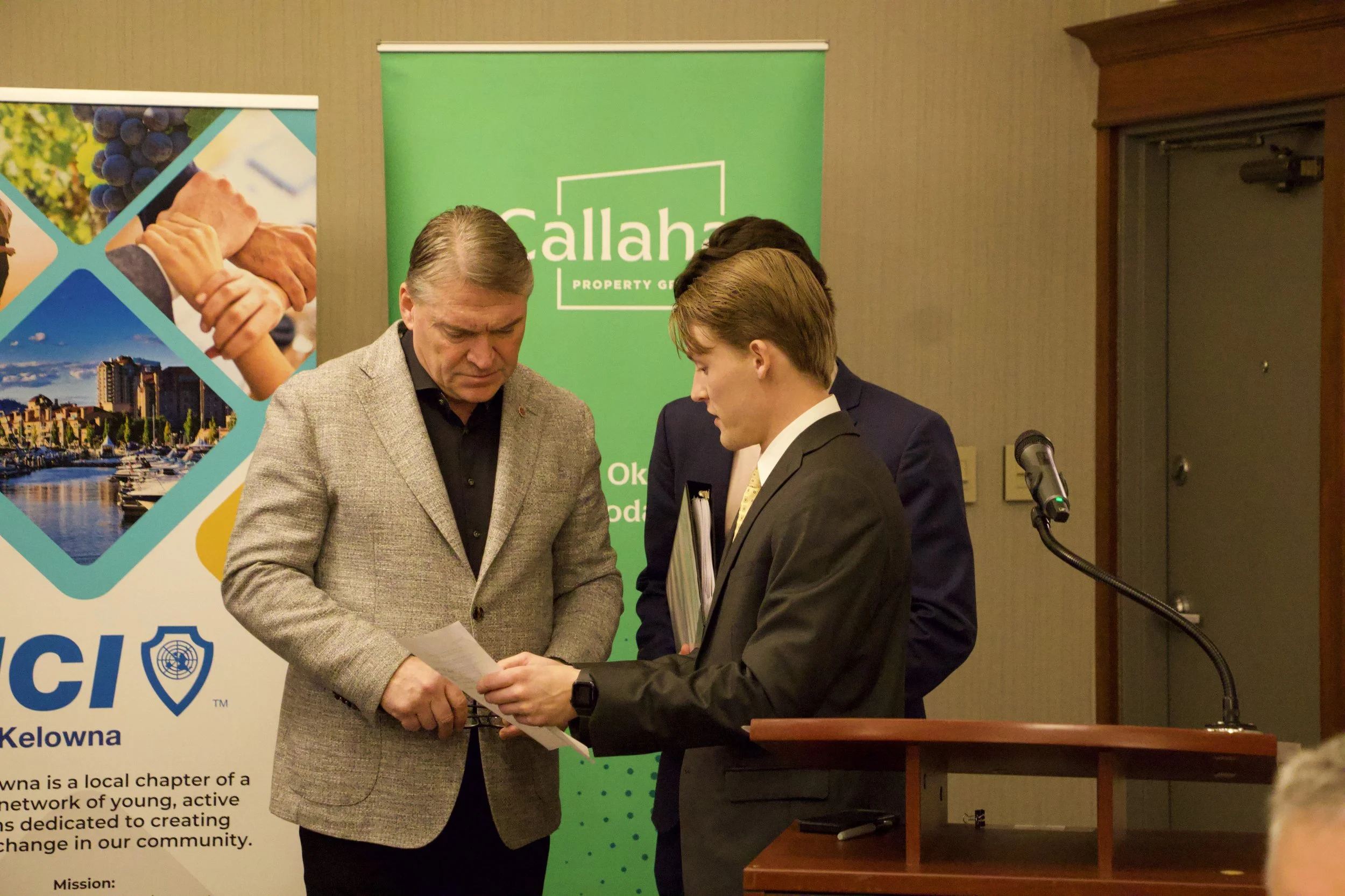 Four men in business attire standing together at a formal event, looking at papers with a microphone and banners in the background.