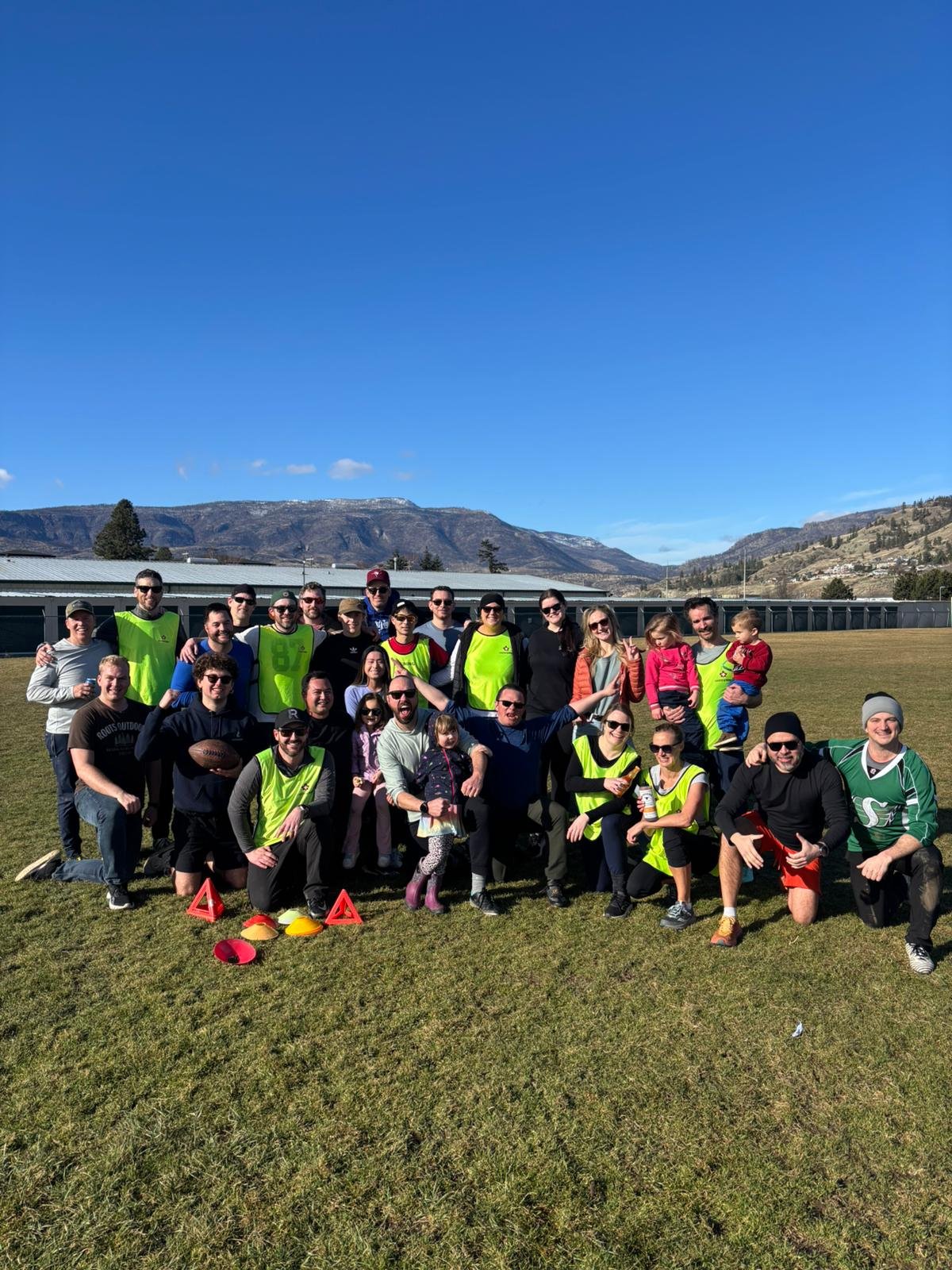 Group of people posing outdoors on a grassy field with mountains in the background, some wearing sports gear.