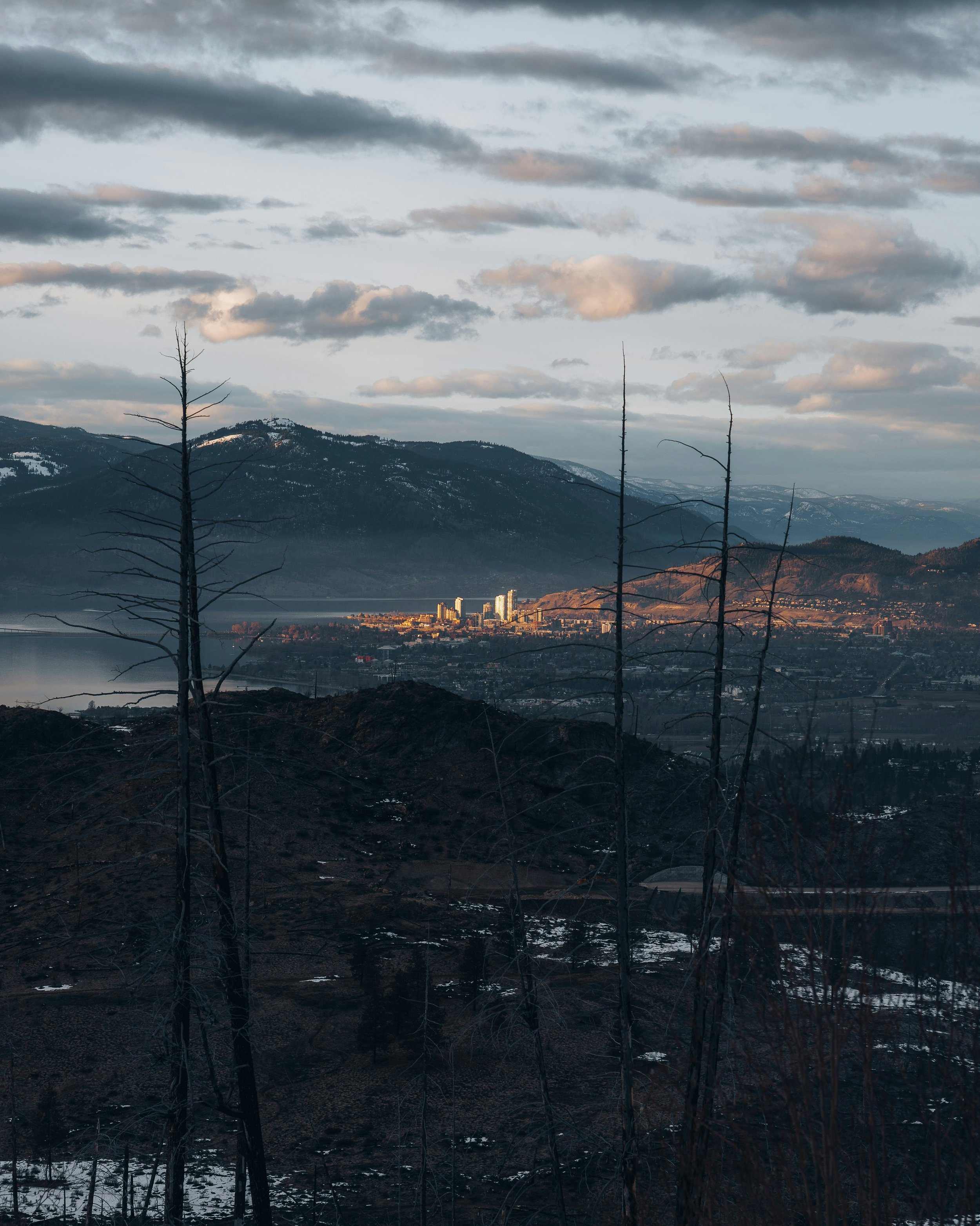 A scenic landscape showing a town near a body of water with mountains in the background, some trees and patches of snow in the foreground, and a cloudy sky.