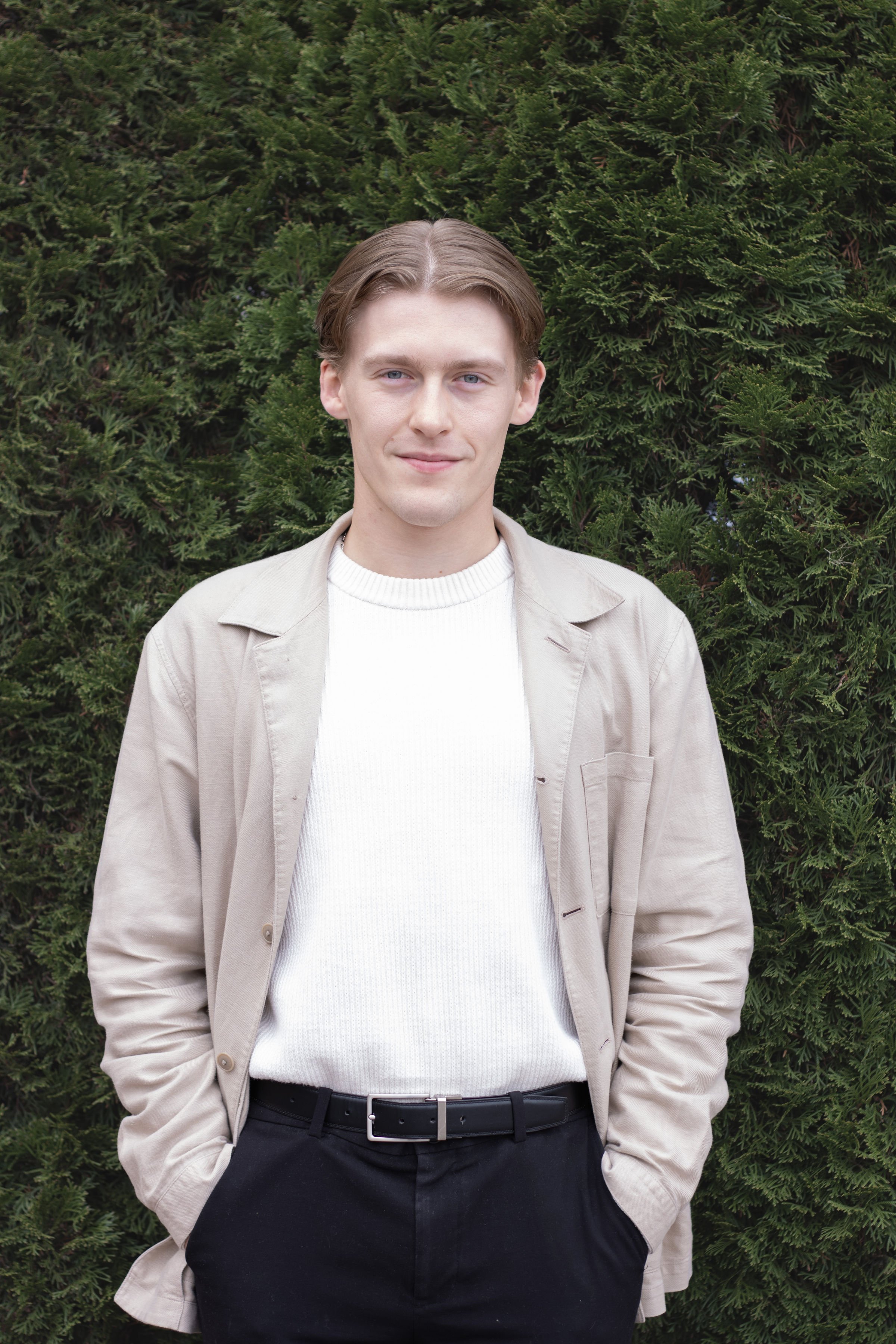 A young man with light brown hair and blue eyes standing outdoors in front of a green hedge, wearing a beige jacket, white sweater, and black pants with his hands in his pockets.