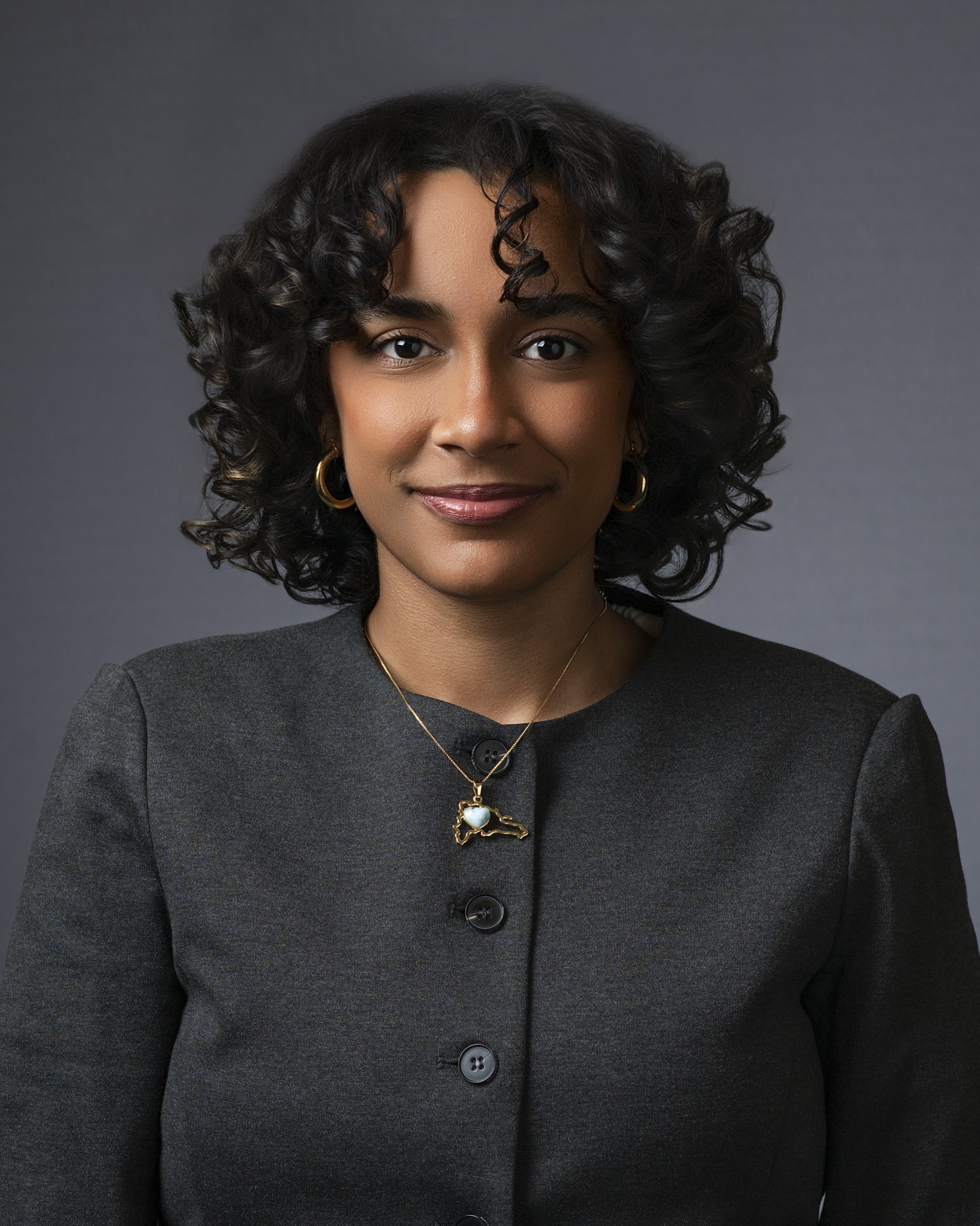 Portrait of a woman with dark curly hair, wearing a dark gray blazer with black buttons, gold hoop earrings, and a gold necklace with a Texas-shaped pendant featuring a blue stone.