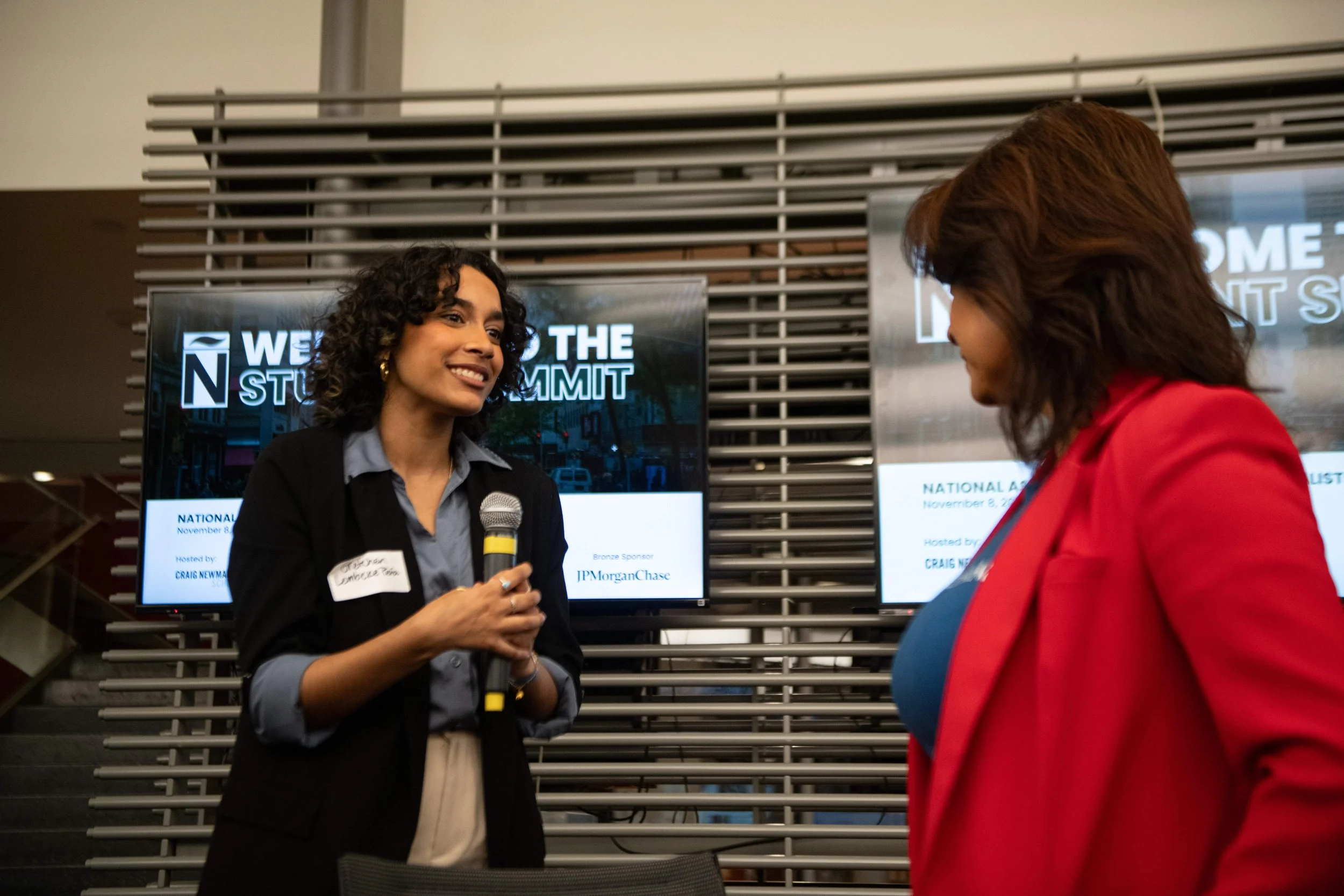 Two women engaged in conversation at a professional event, with digital screens in the background displaying the event's title and details.