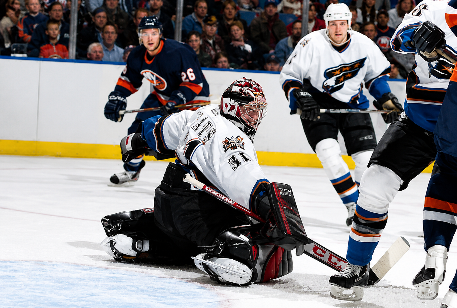 An ice hockey game where players are around the goal, with a goalie in black kneeling on the ice attempting to block a shot. Spectators are visible in the background.
