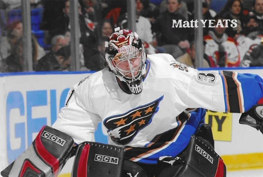 Hockey goalie in white jersey with a colorful logo, wearing protective gear, in action during a game with fans in the background.
