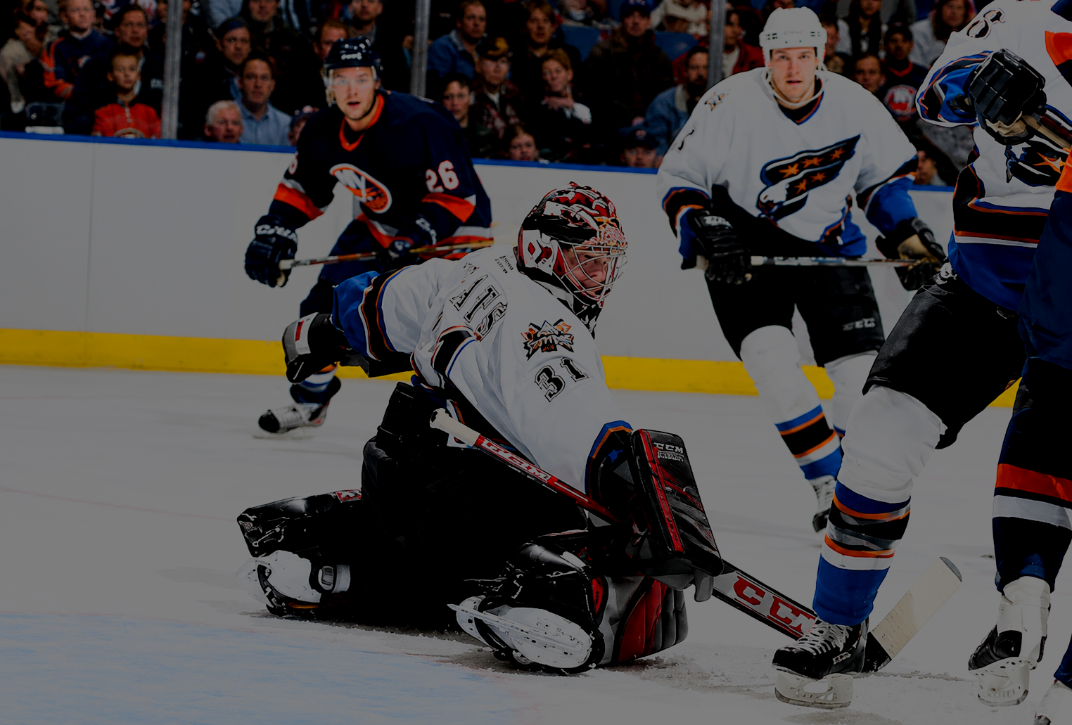 Hockey players during a game with a goalie in white uniform making a save, and other players in team jerseys around him, with spectators watching in the background.