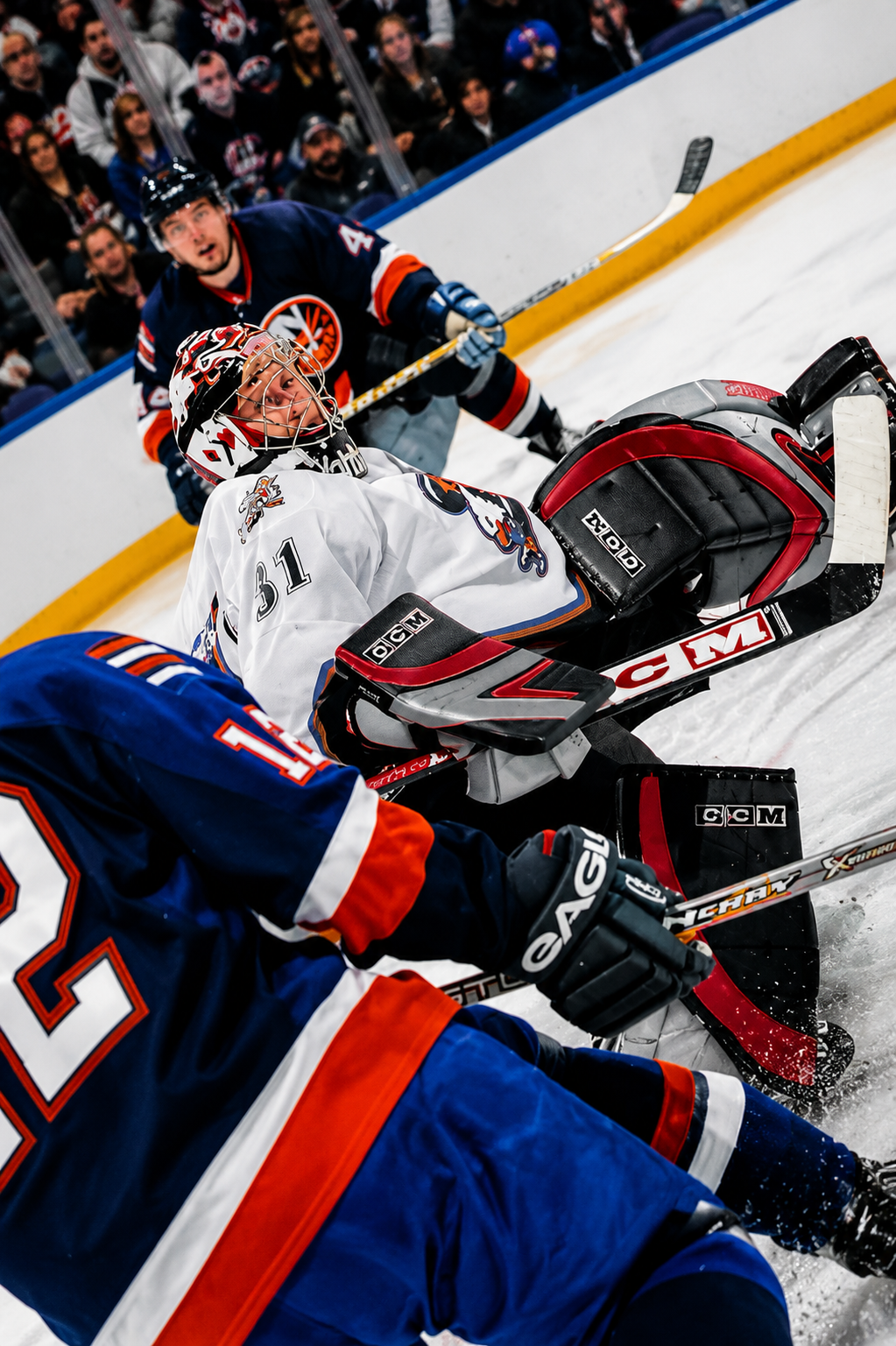 Hockey game scene with a goalie in white gear on the ice, surrounded by players in blue and red jerseys, and spectators watching behind the glass.