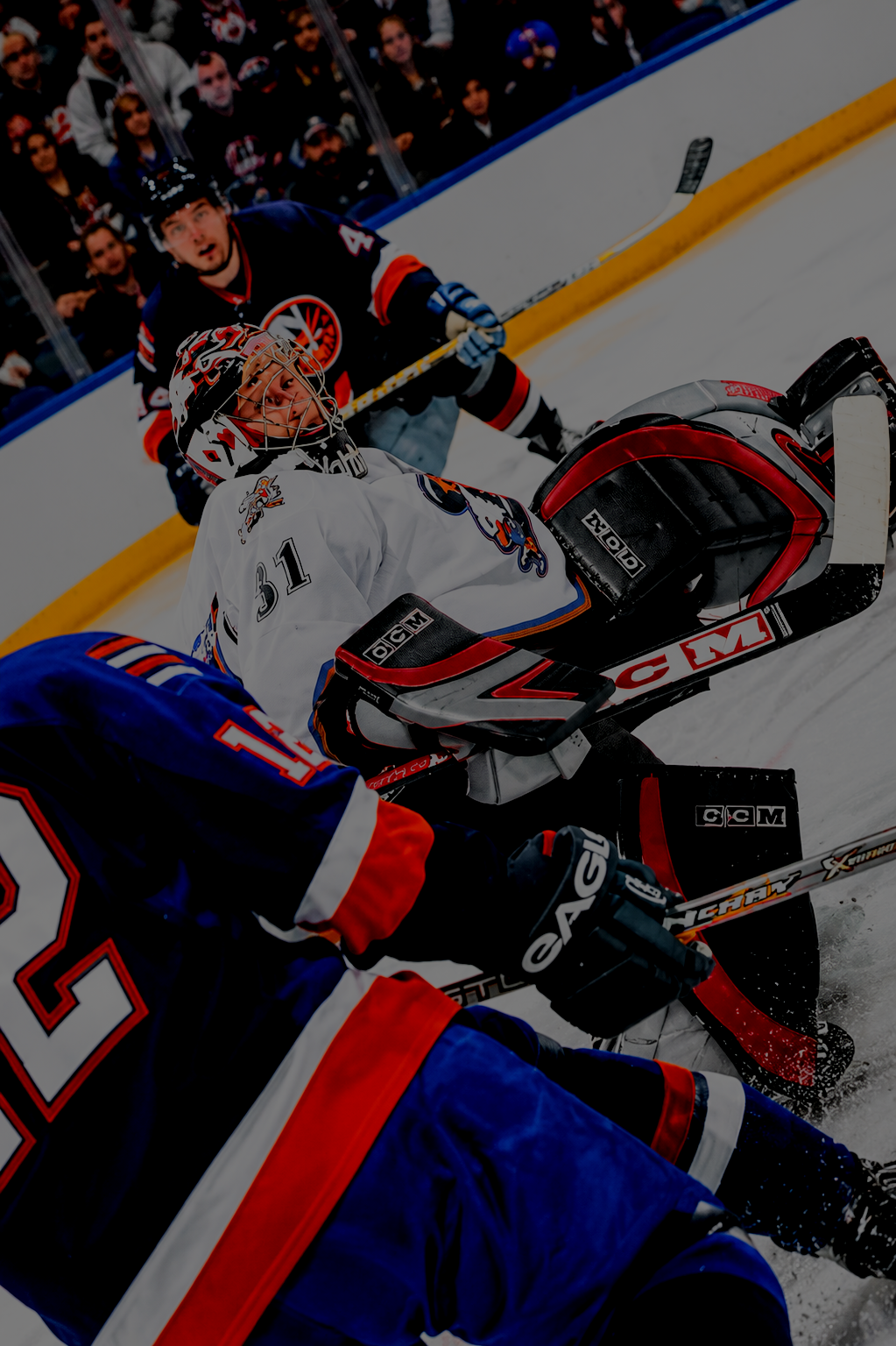 Hockey players during a game, with one goalie in white gear and two skaters in blue and red jerseys, on ice rink with spectators watching.