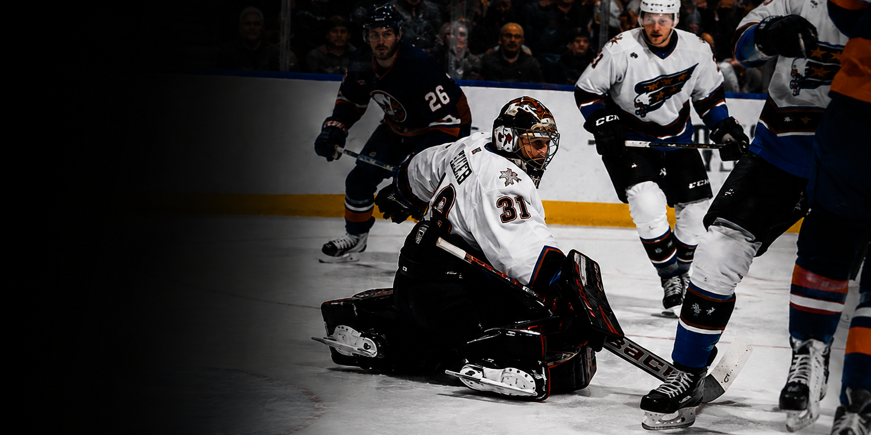 Ice hockey players in action during a game, with one player in white goalie gear kneeling on the ice and others in dark and white jerseys nearby.