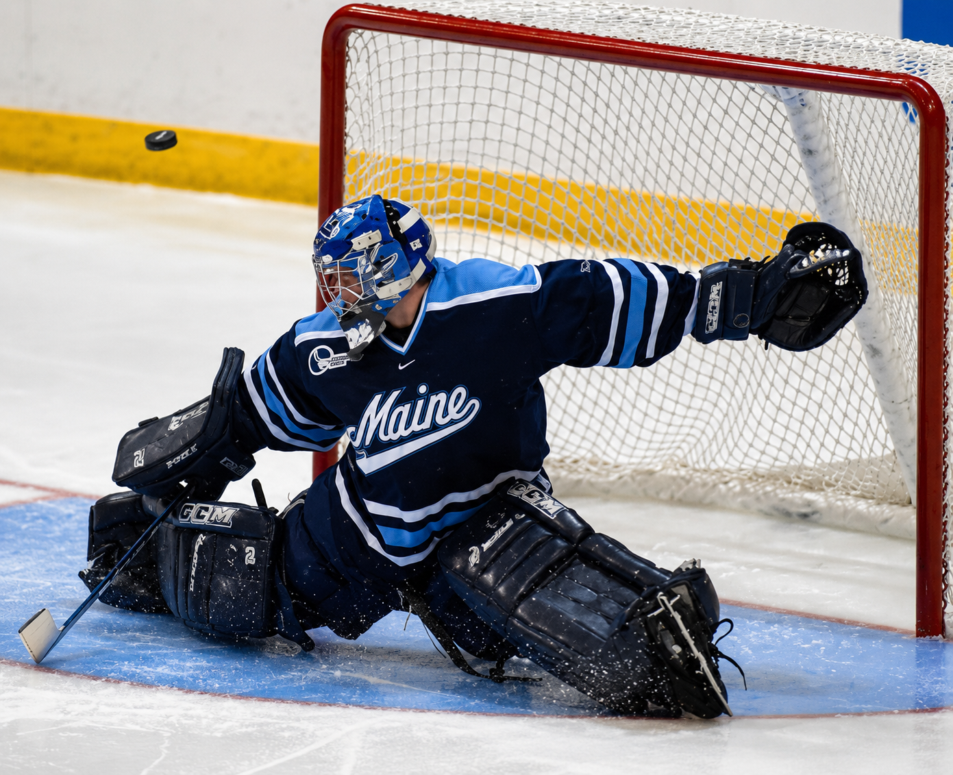 An ice hockey goalie in a blue Maine team uniform, kneeling on the ice, blocking the goal with their glove and leg pads.