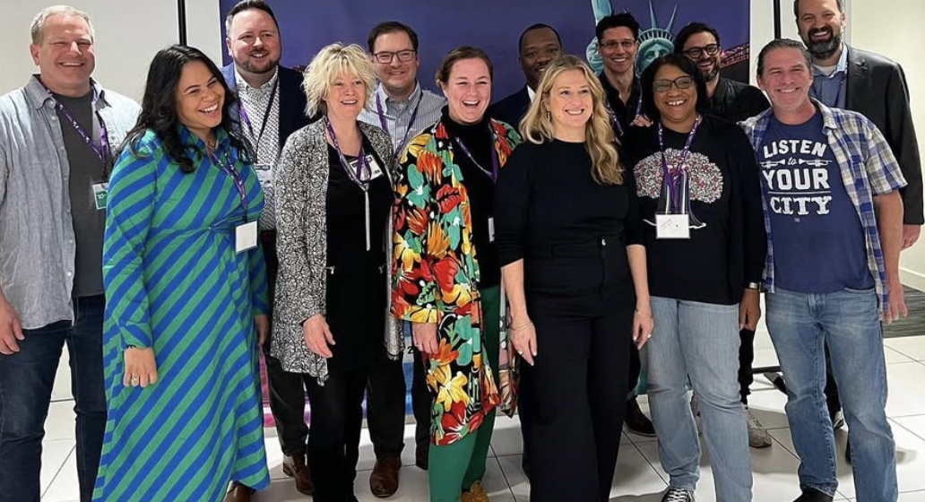 A diverse group of twelve people, smiling, gathered indoors for a group photo. Some are wearing conference lanyards, and a backdrop with a purple and blue design is visible behind them.