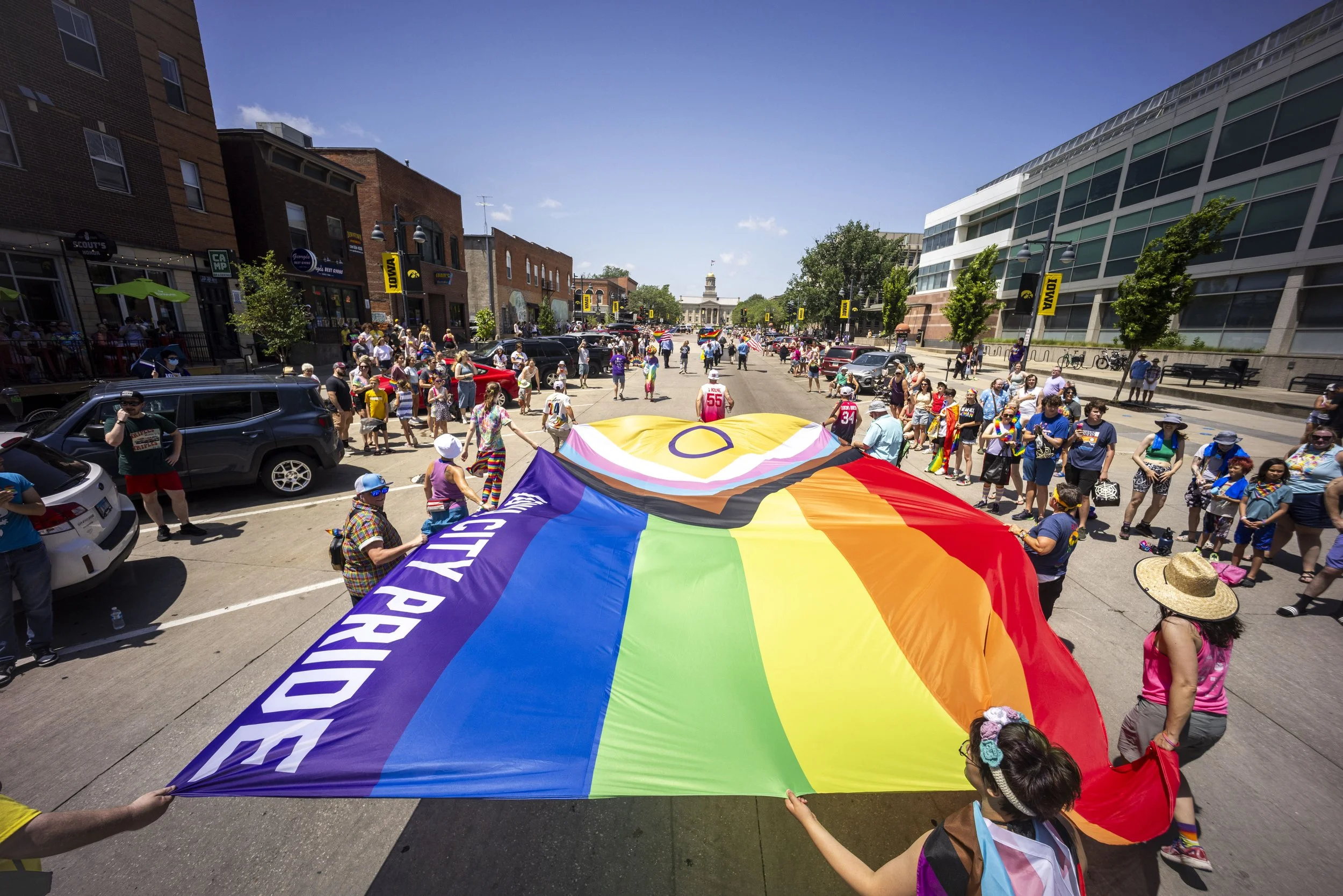People participating in a Pride parade on a city street, holding a large rainbow Pride flag with the words 'City Pride' on it, surrounded by spectators and buildings.