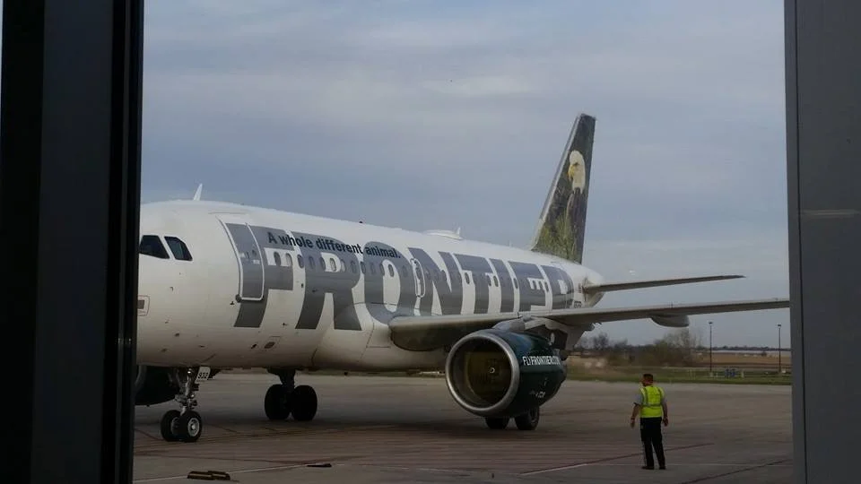 An airplane on the tarmac with the word 'FRONTIER' on the side, a yellow-vested worker standing nearby, and a sky with grey clouds.