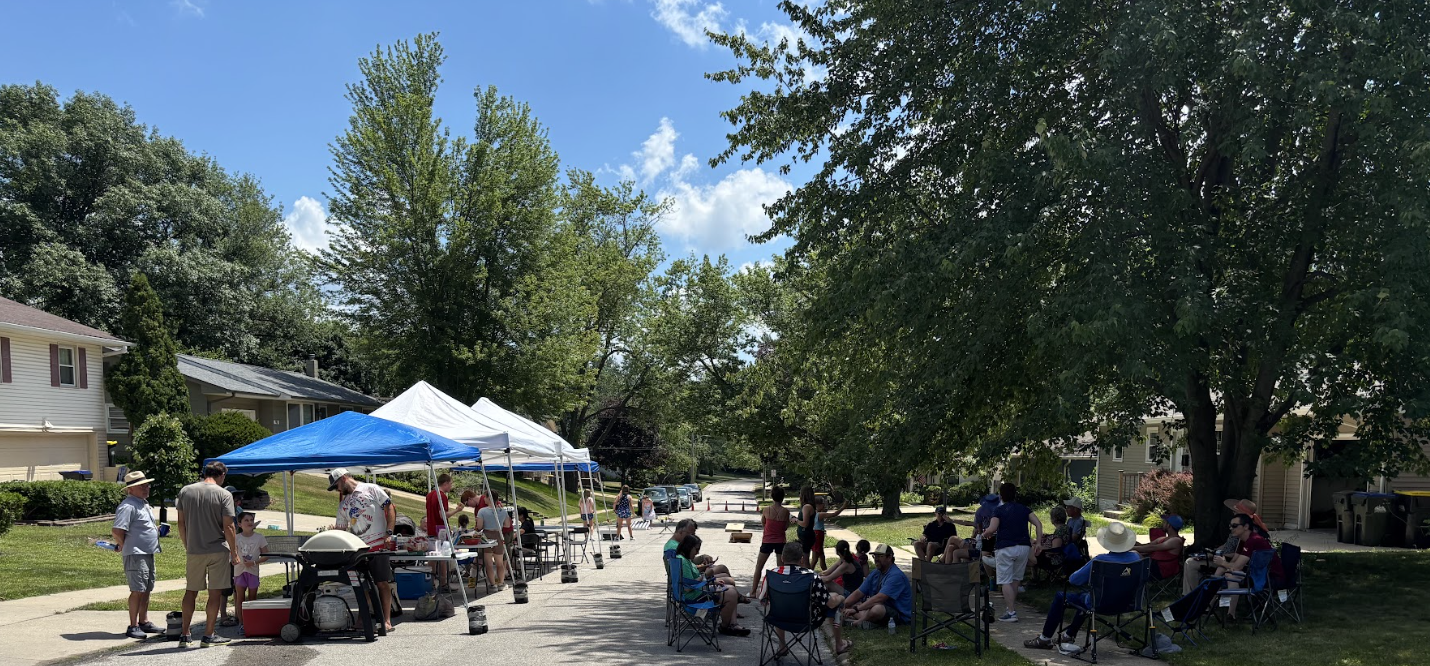 Community gathering on a residential street with tents, tables, and people sitting and standing in the shade of trees on a sunny day.