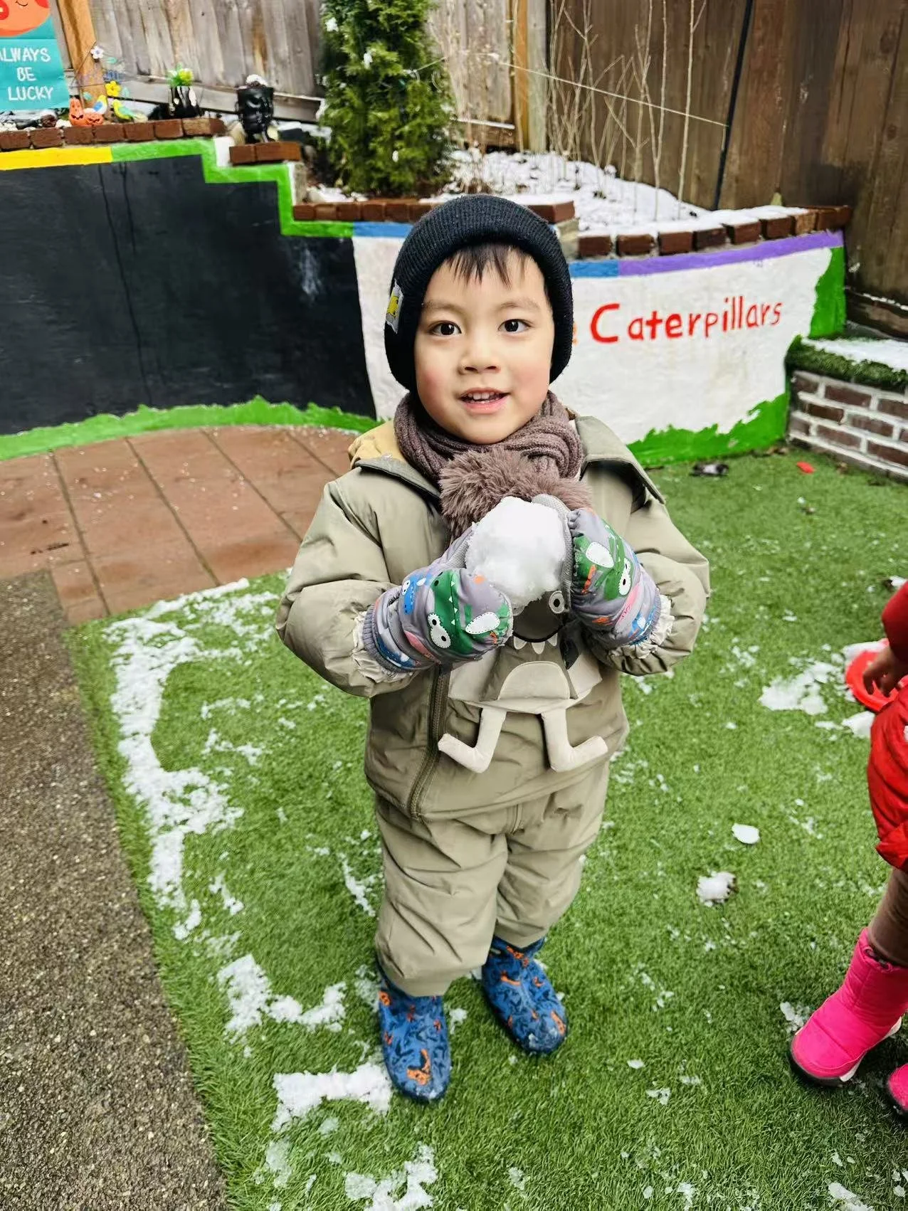 A smiling child in winter clothing holds a snowball while standing on a lightly snow-covered playground.