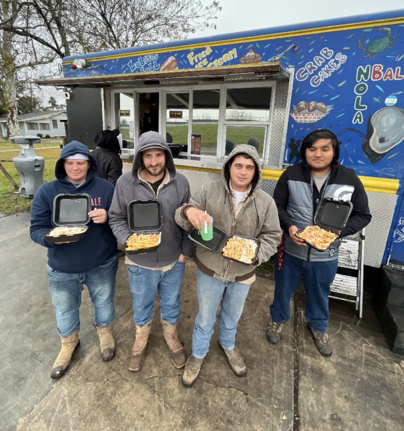 Four people standing in front of a food truck, each holding a takeout container of food, with two of them wearing hoodies and rain boots, under an overcast sky.
