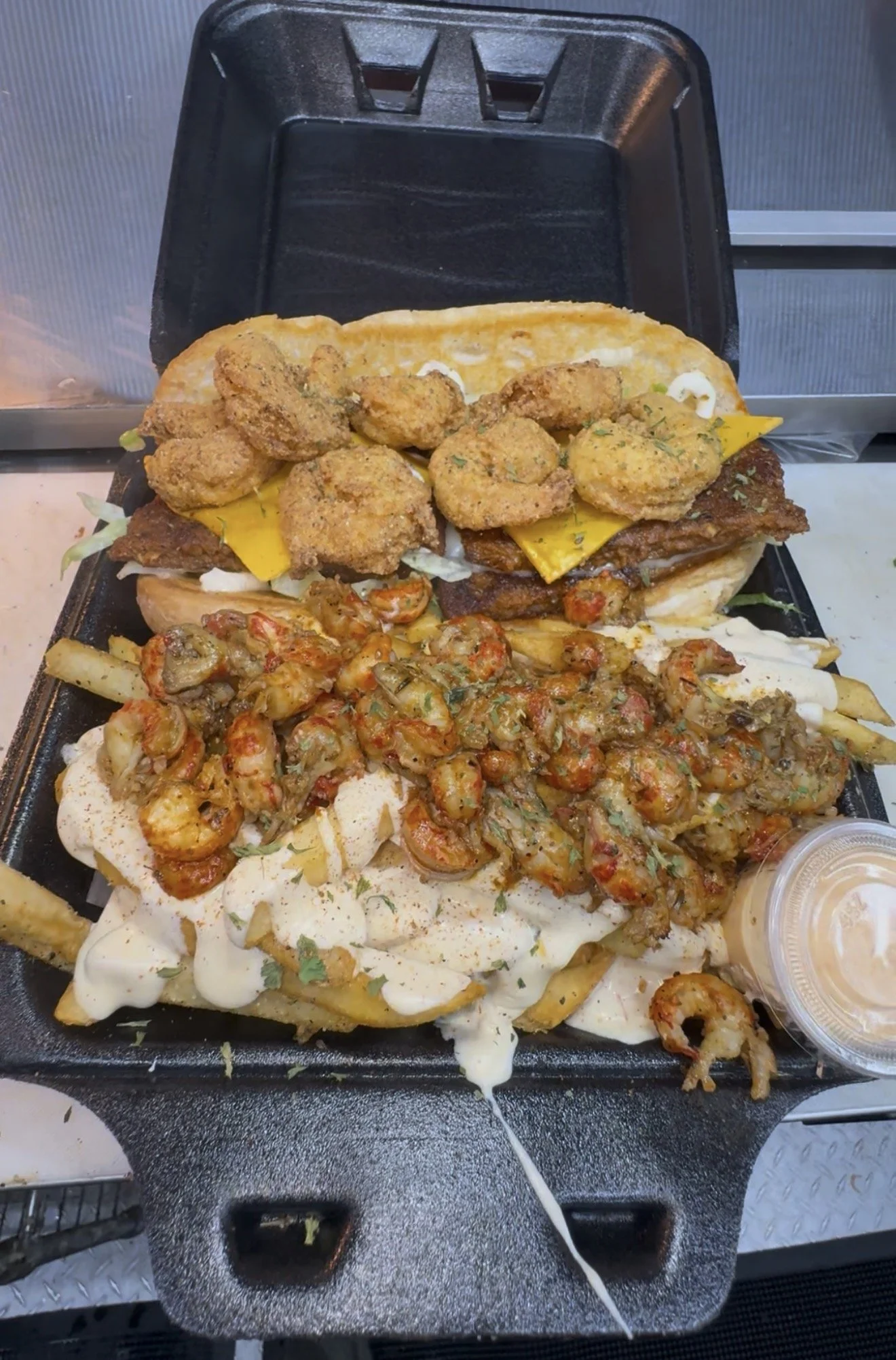Loaded takeout dinner with fried shrimp, fried fish, fries, cheese fries, shrimp topping, and dipping sauce in a foam container.