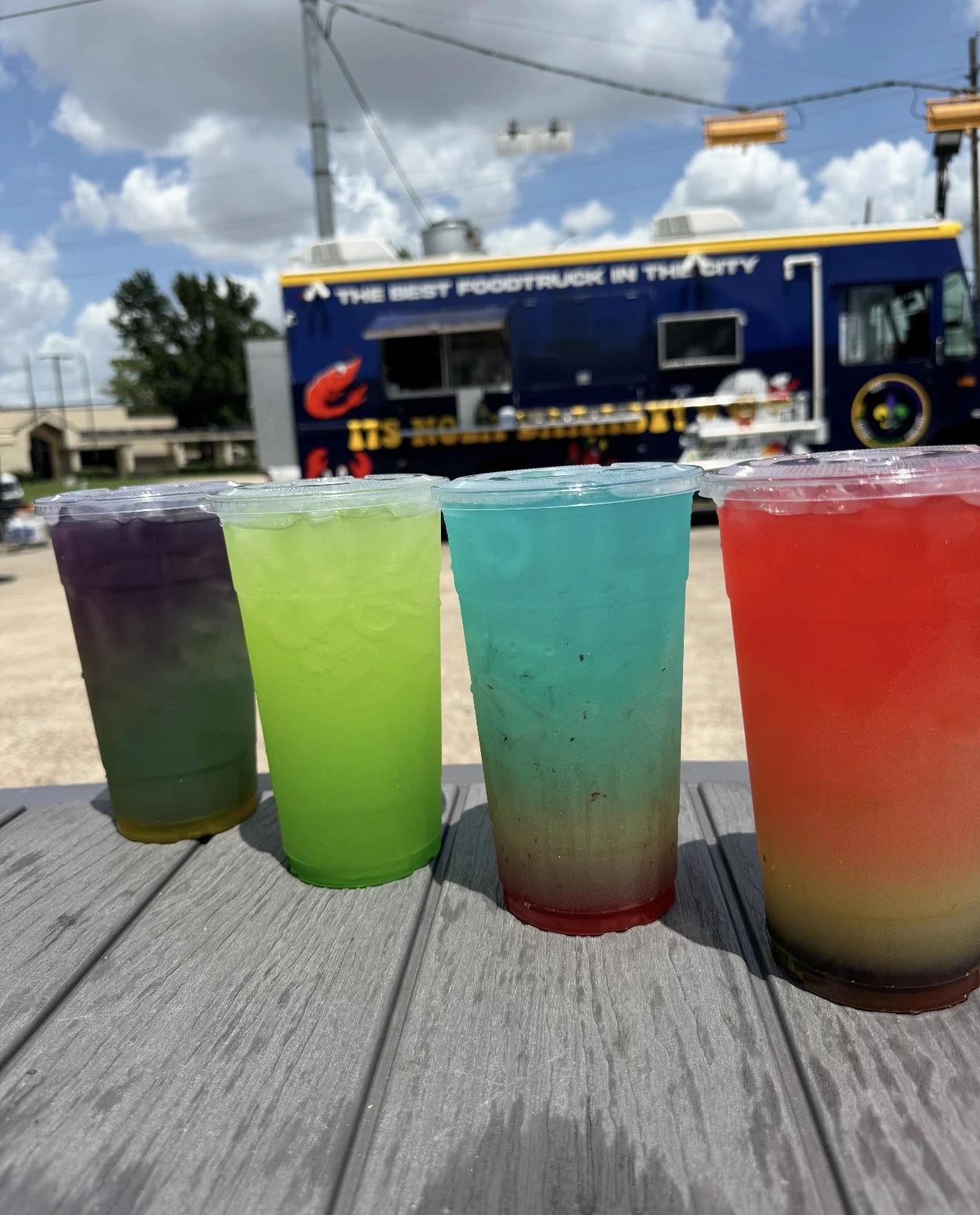 Four colorful twisted lemonade drinks  in plastic cups with a food truck in the background under a partly cloudy sky.