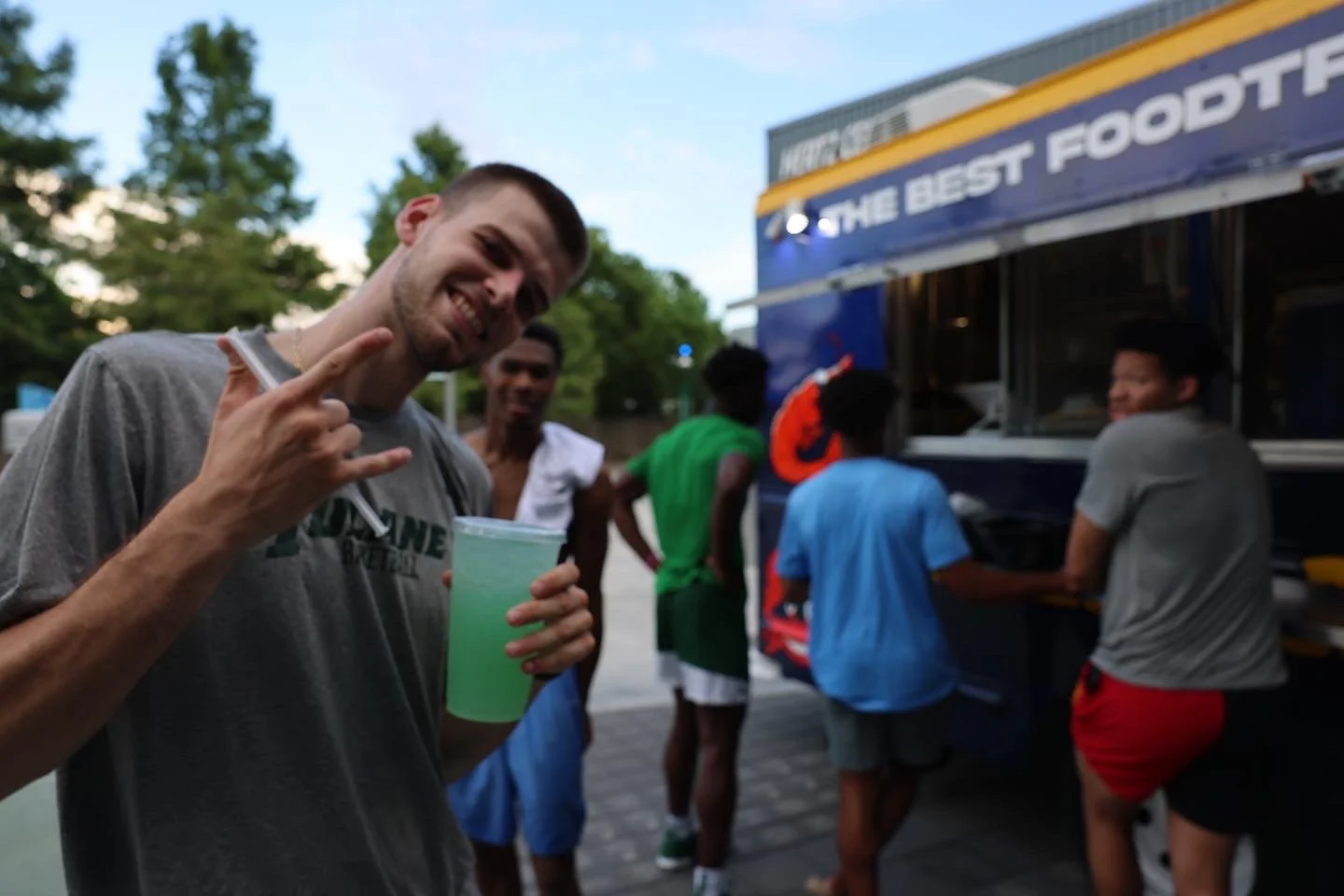 A smiling young man in a gray T-shirt making a rock and roll hand gesture while holding a green drink in front of a food truck with several people gathered around.
