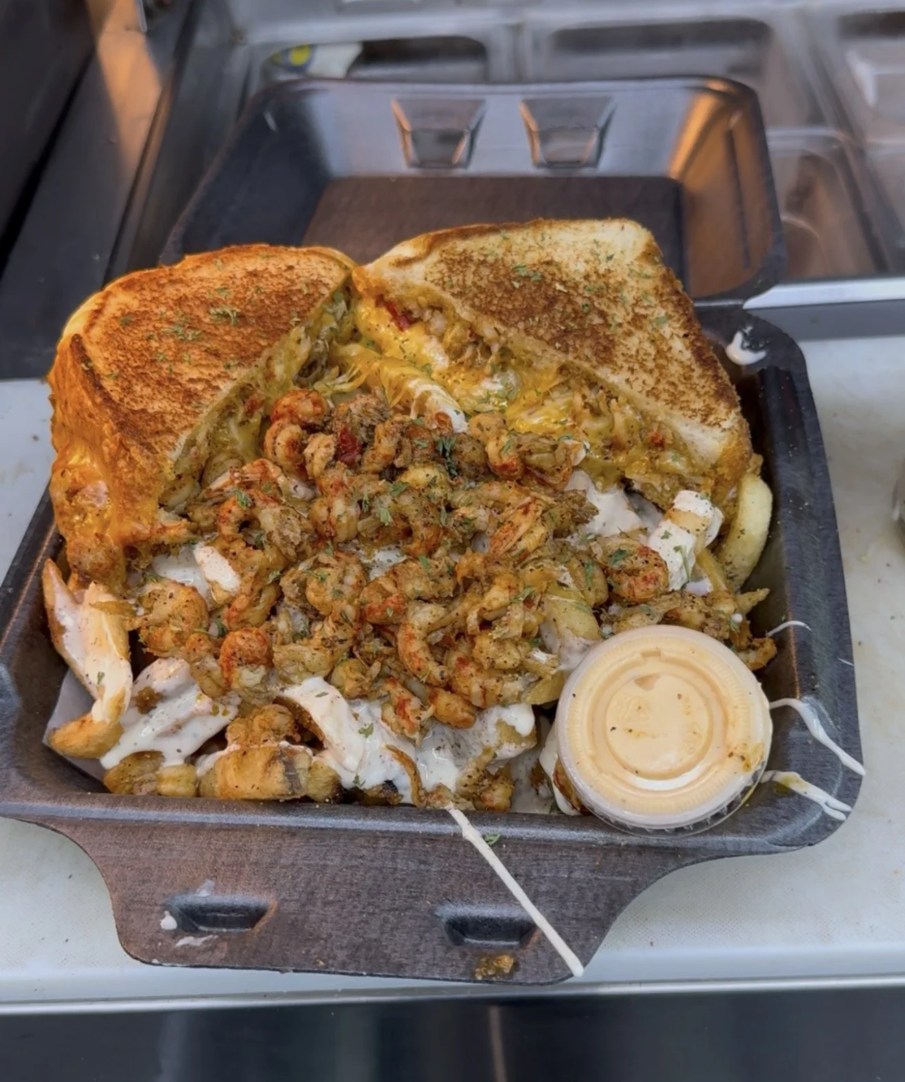 A tray of street food featuring a grilled sandwich with fried chicken, chopped fried shrimp, shredded lettuce, mayonnaise, and a small container of mustard or sauce.