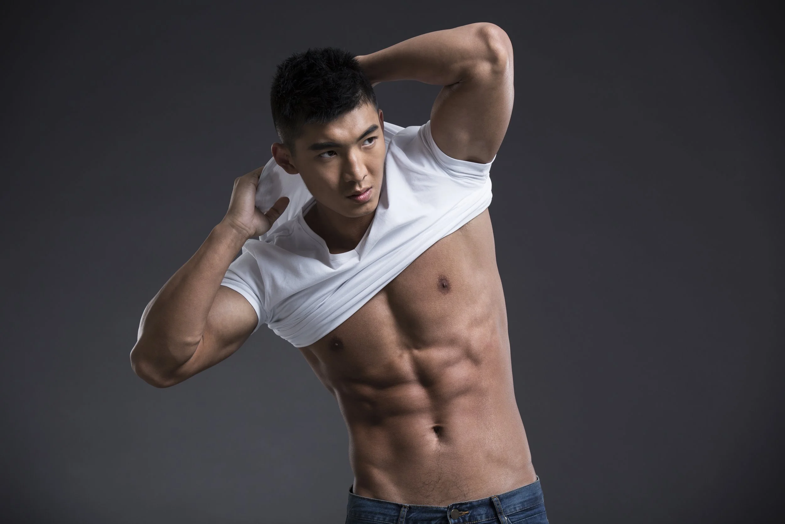 Young man with a muscular physique, wearing jeans and a white t-shirt, lifting his shirt to reveal abs against a dark background.