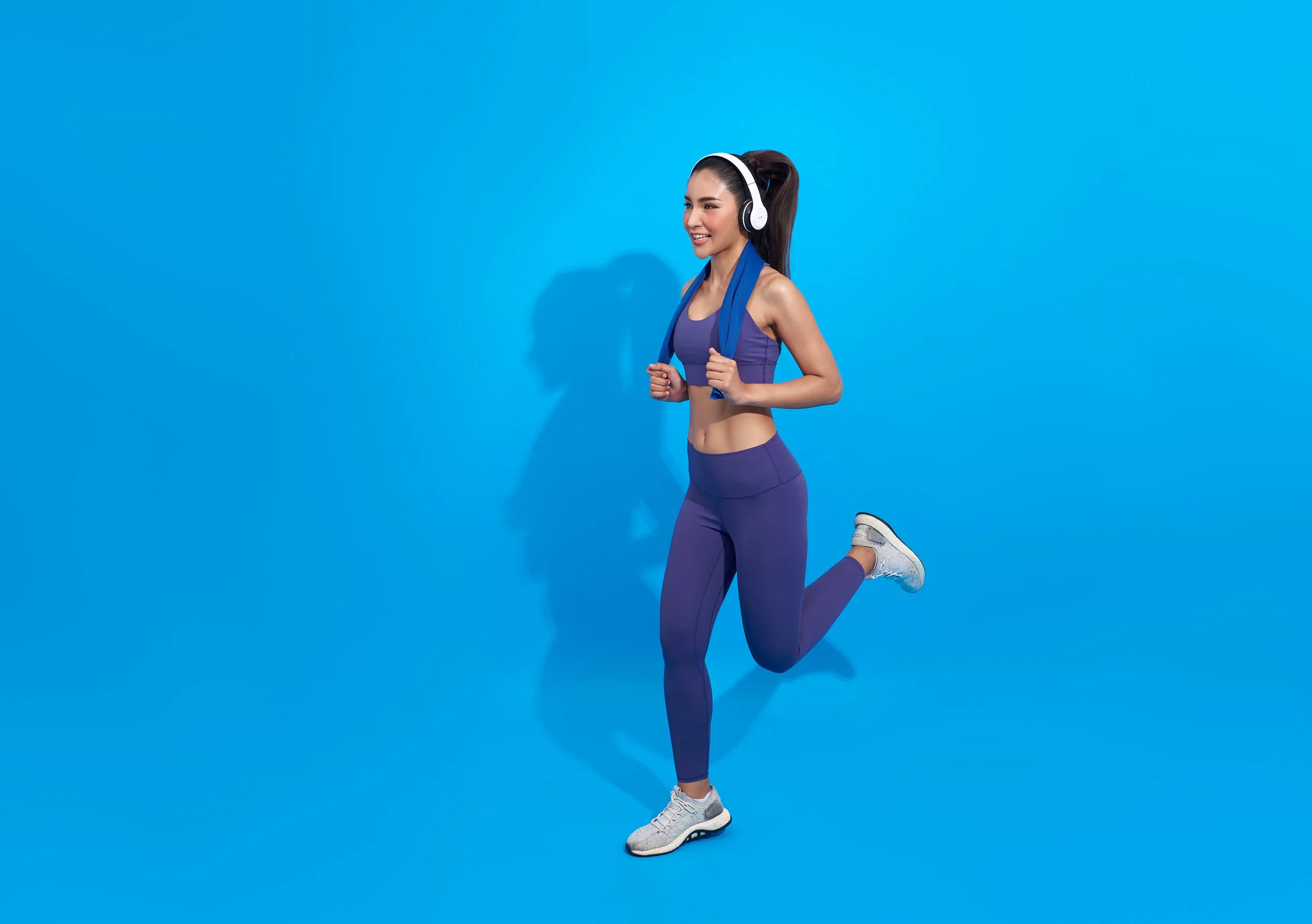 Young woman in purple workout clothes, wearing headphones, with a blue towel around her neck, jogging against a blue background.