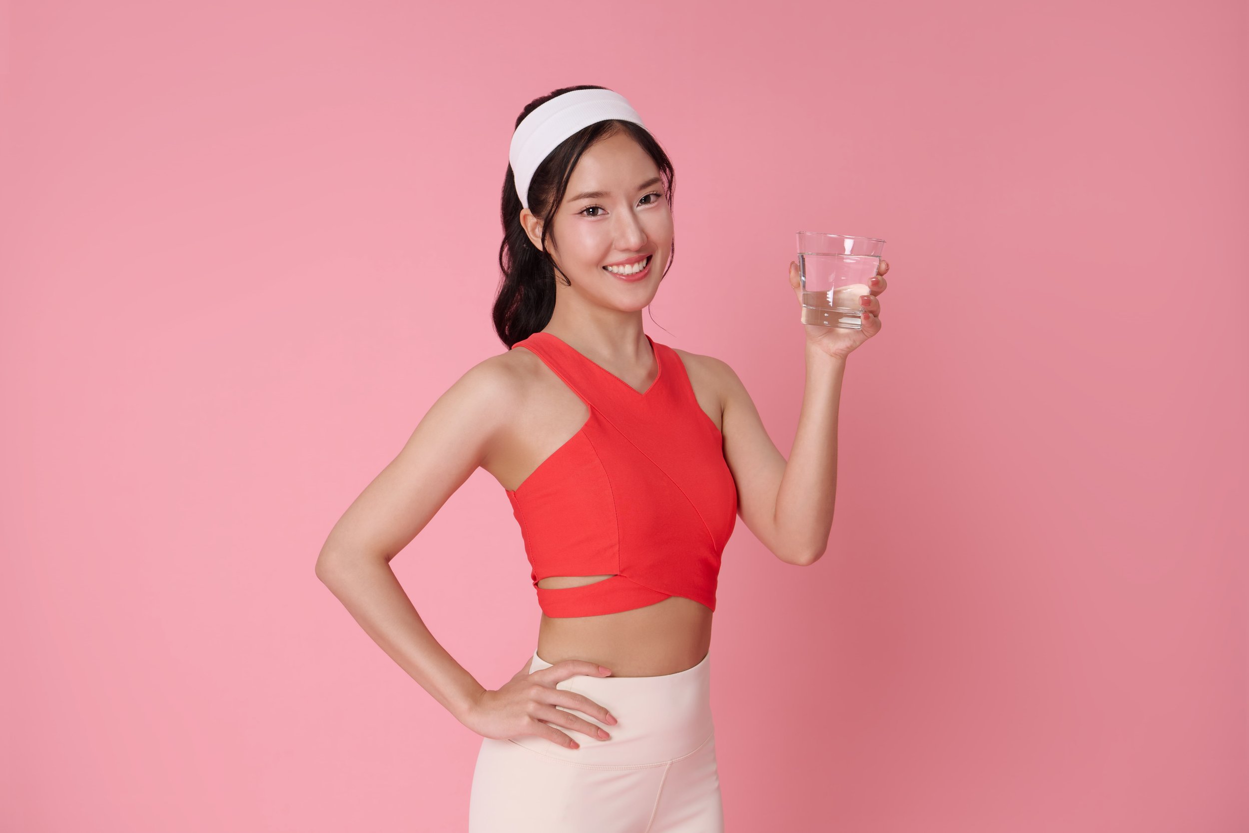 A young woman in a red top and beige pants holding a glass of water, smiling, standing against a pink background.