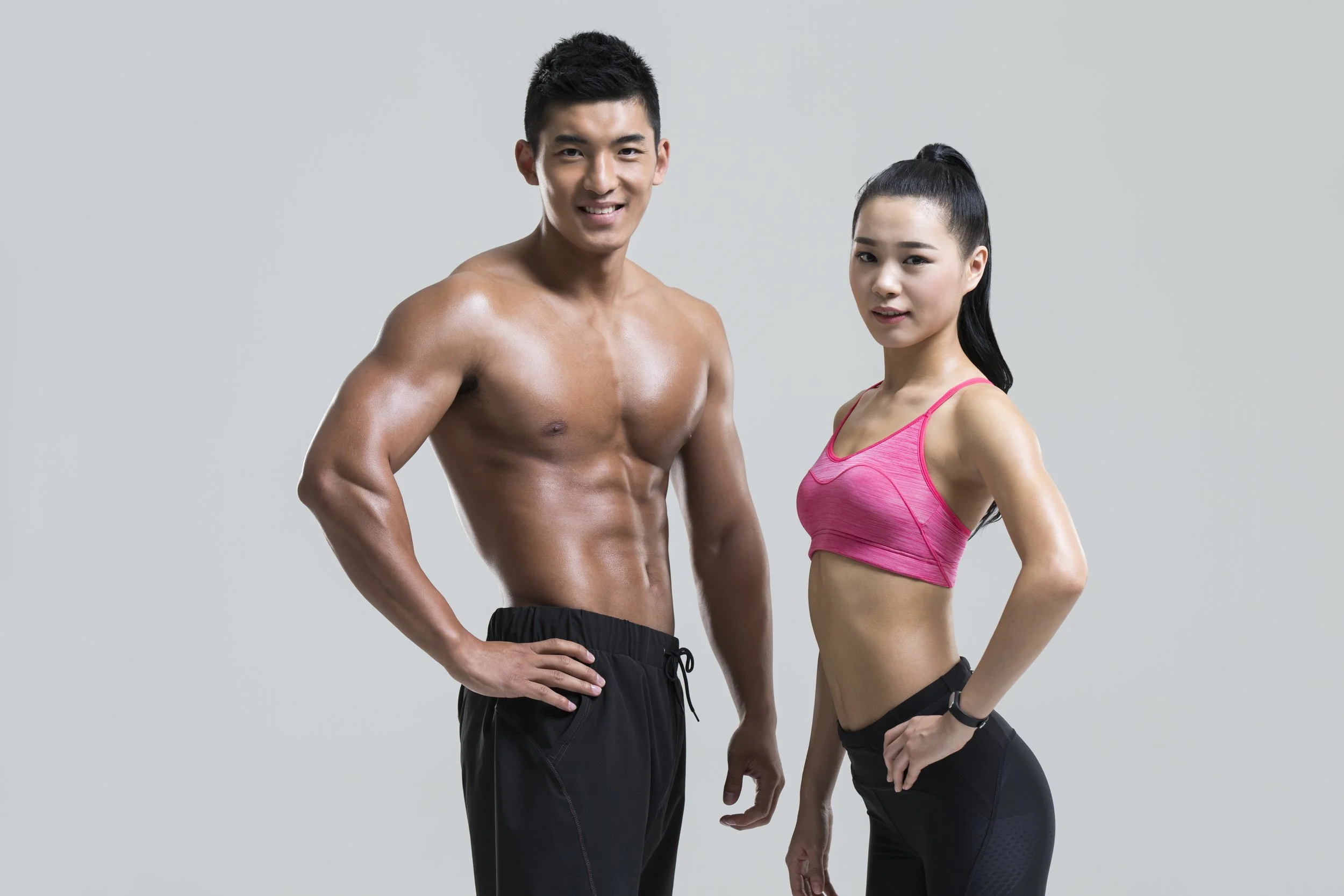 A fit man and woman in workout attire standing against a gray background, smiling and posing.