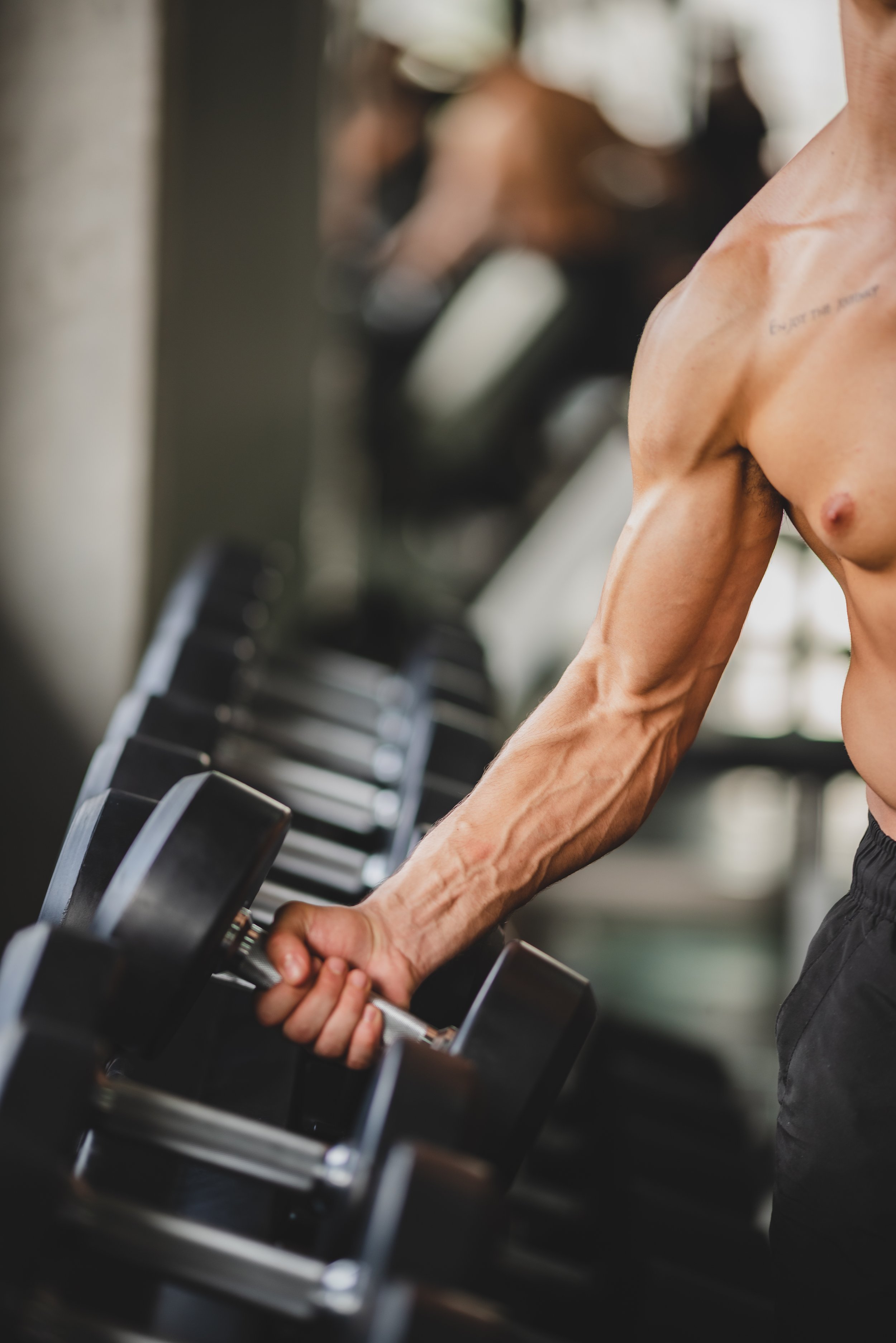 Close-up of a muscular man lifting a dumbbell in a gym, with a row of dumbbells in the background.