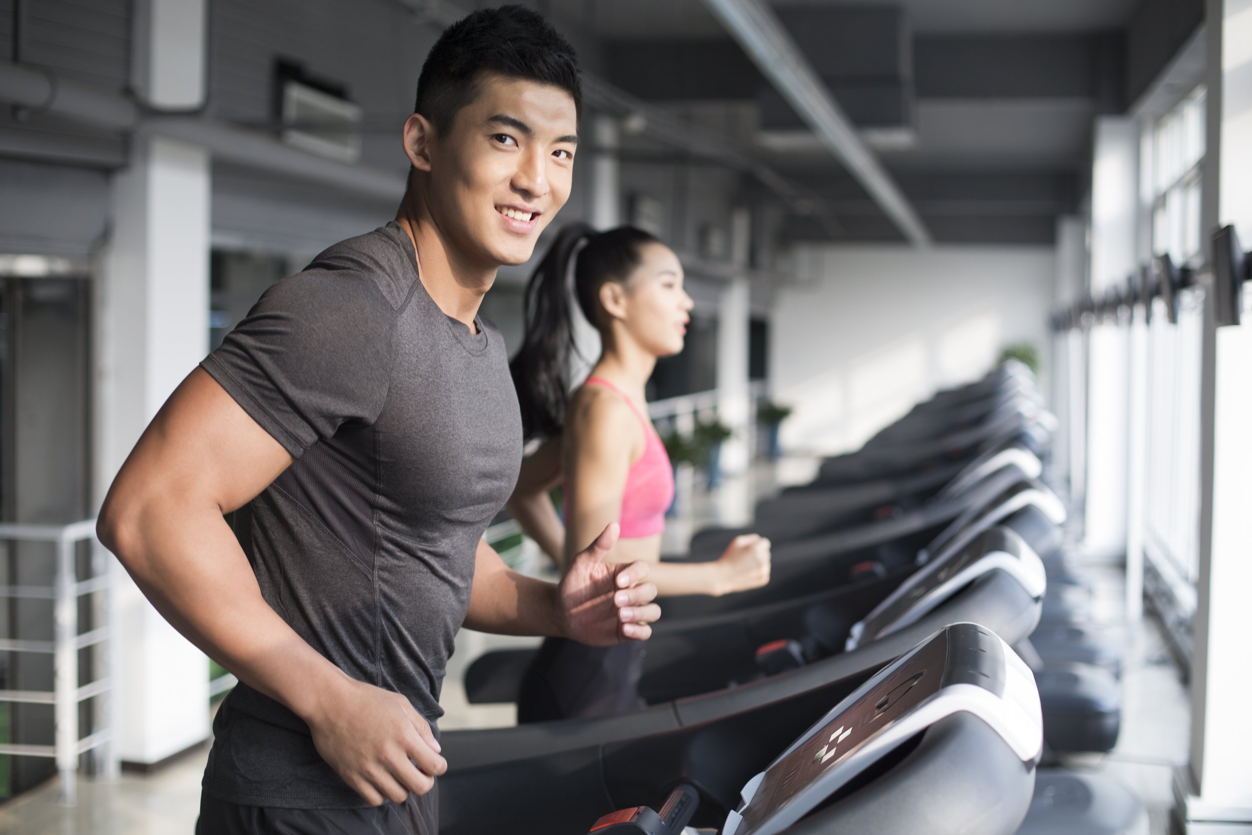 Two people exercising on treadmills in a modern gym with large windows, a man in the foreground smiling at the camera and a woman in the background in pink workout attire.