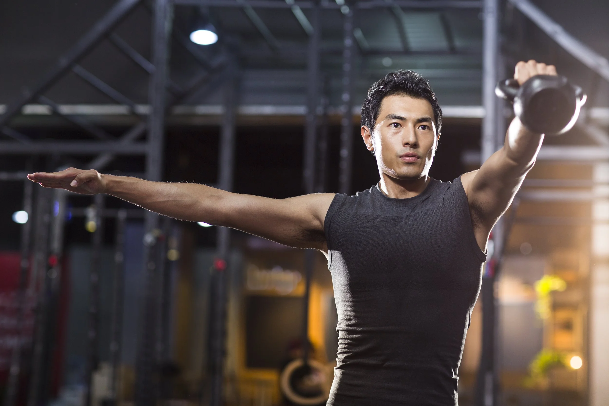 A man lifting a kettlebell in a gym, extending his right arm forward and his left arm to the side, with gym equipment in the background.