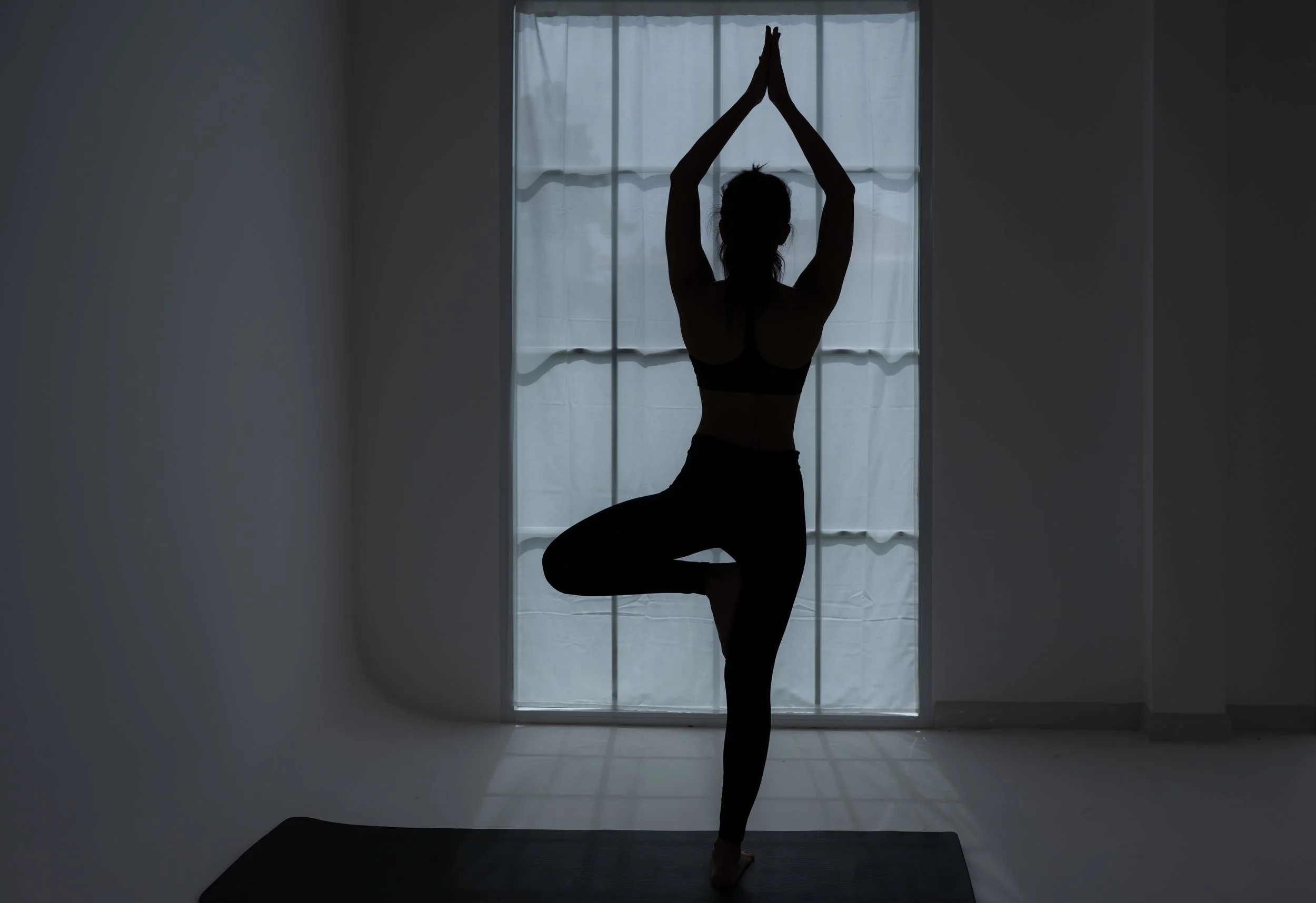 Silhouette of a woman practicing yoga indoors near a window, standing on one leg with hands pressed together above her head in the tree pose.