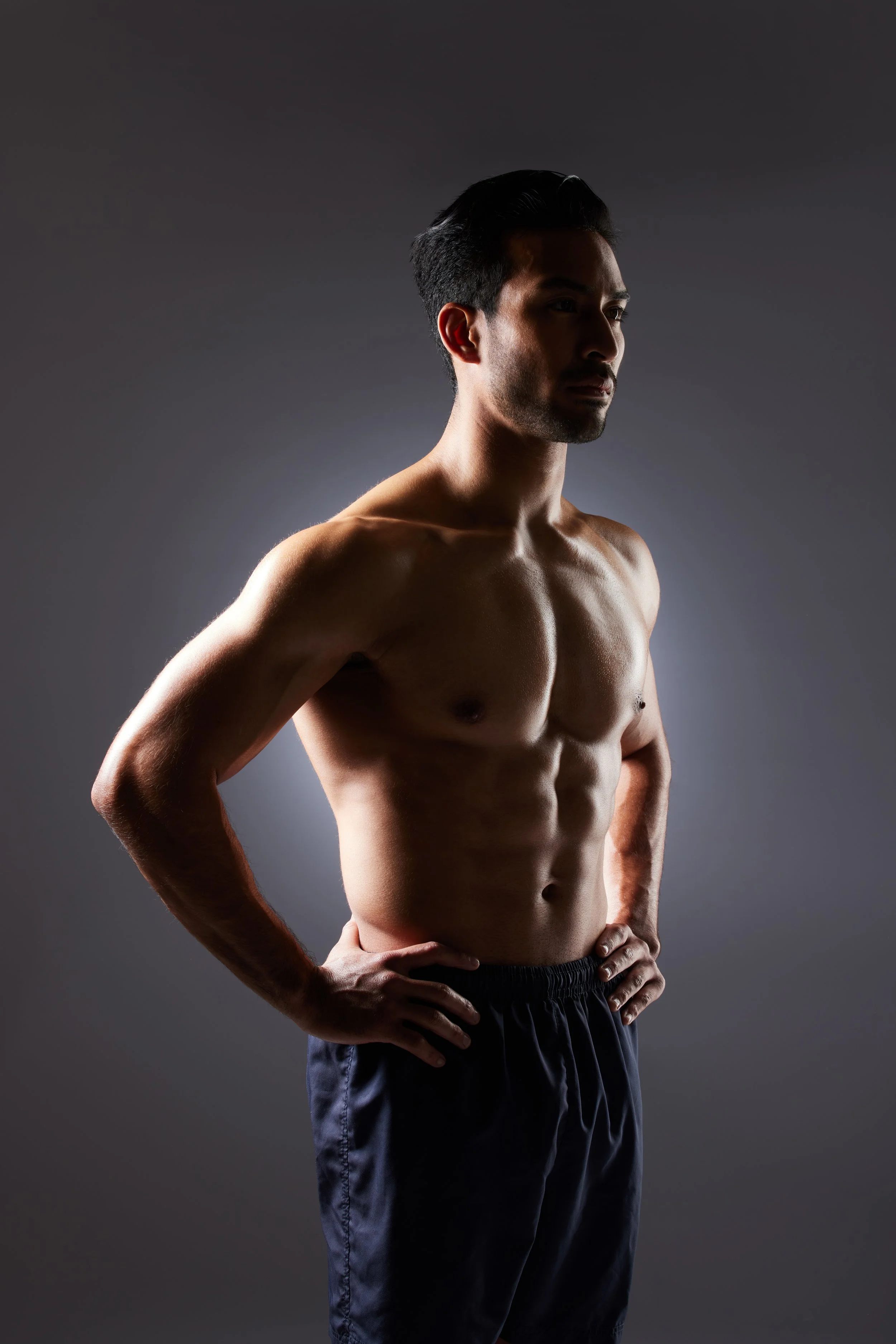 A shirtless man with hands on hips, standing against a dark background, looking to the side.