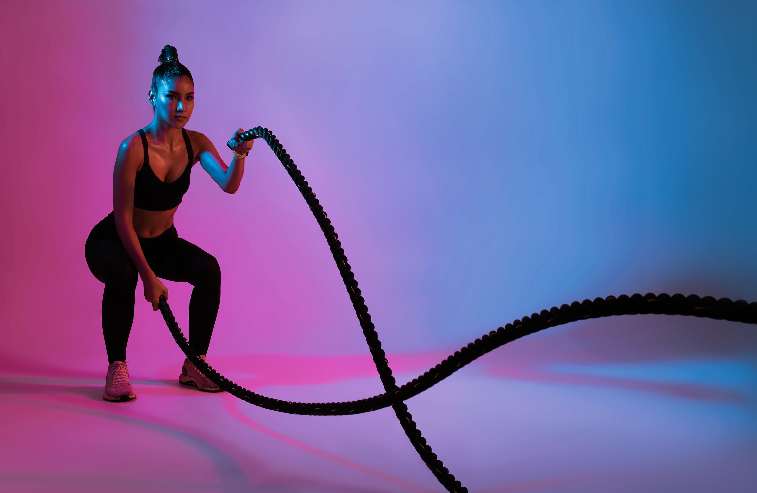 A young woman in workout attire, holding battle ropes in a workout studio with colorful pink and blue lighting.
