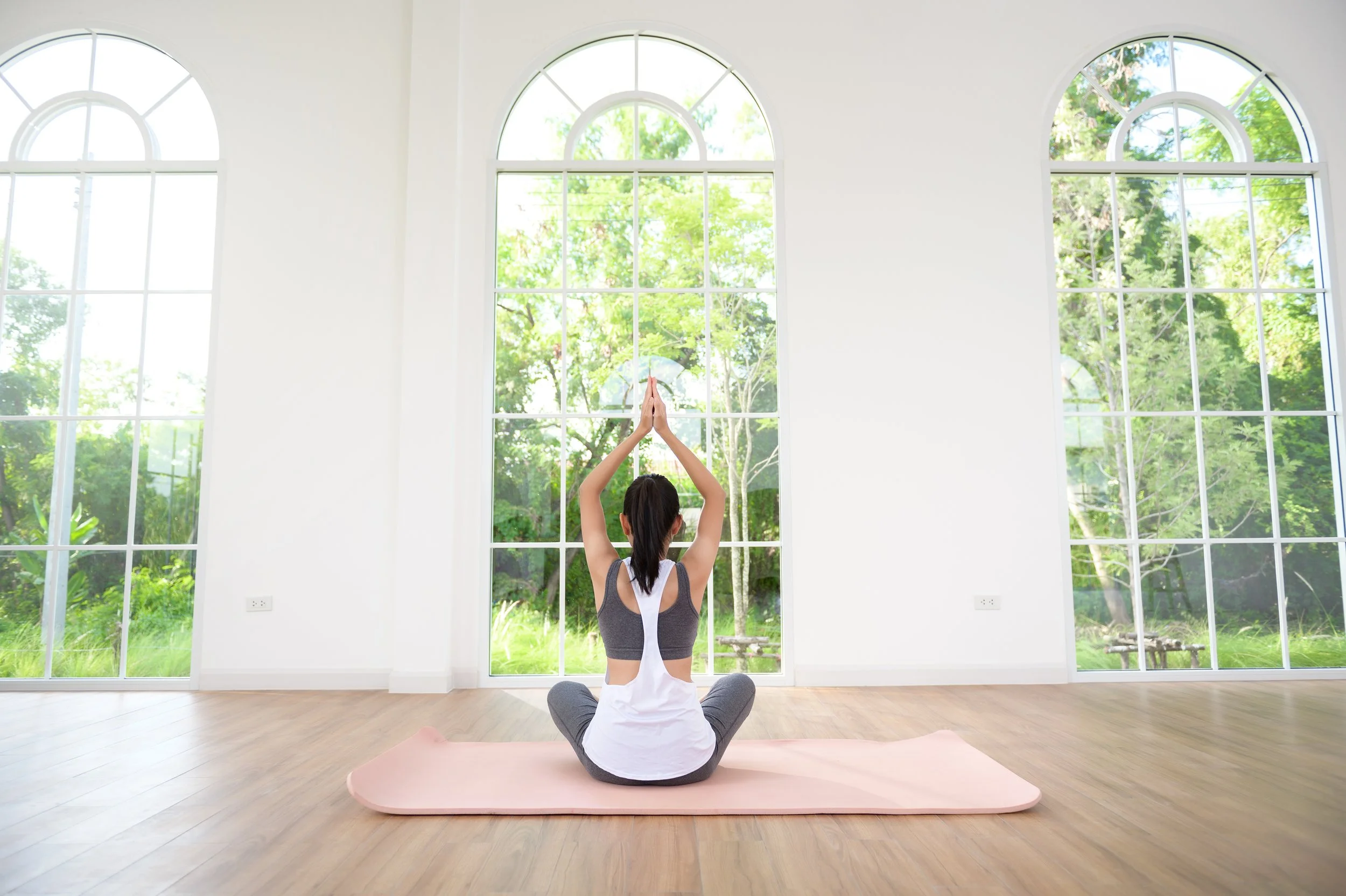 A woman practicing yoga in a bright, spacious room with large arched windows showing green trees outside.