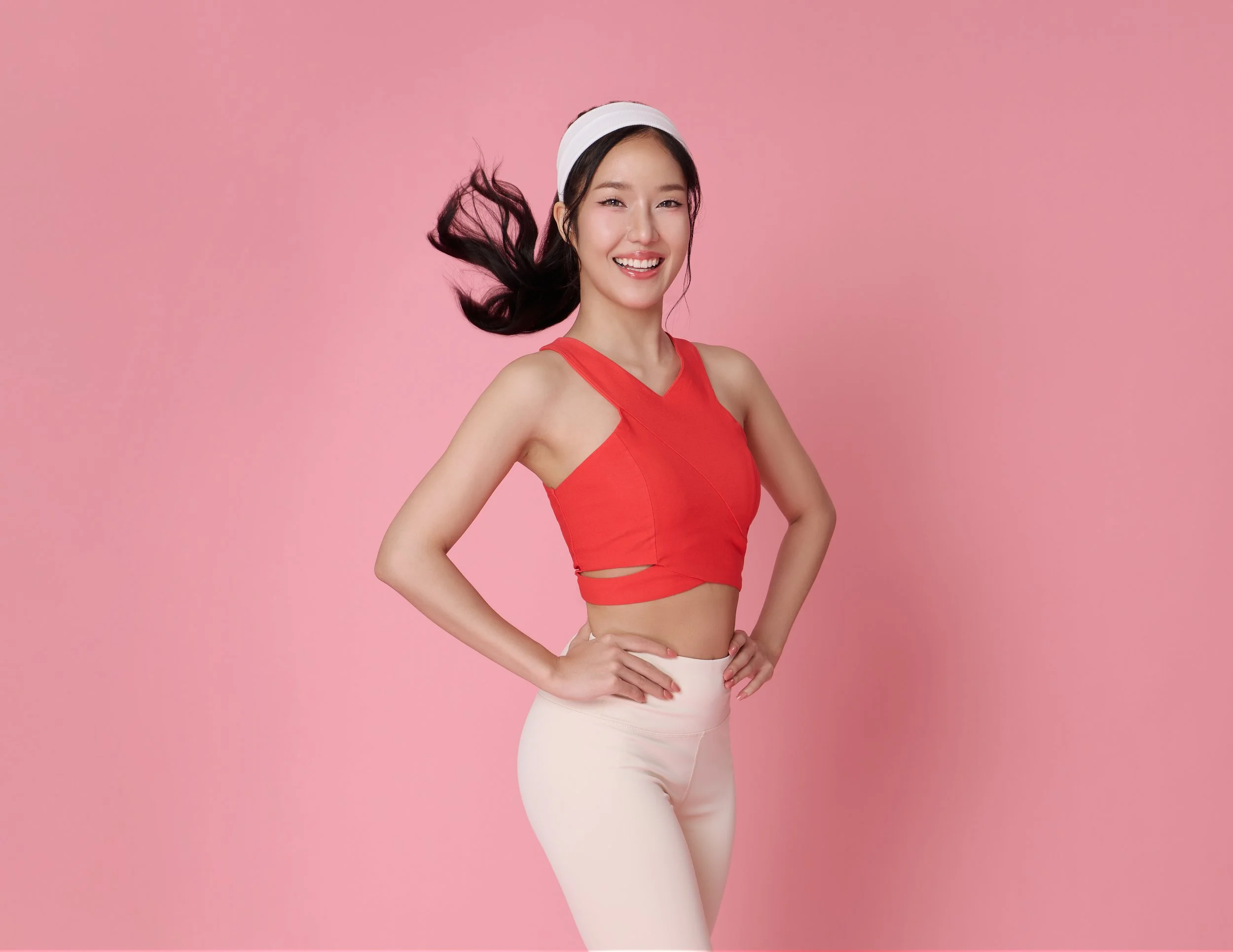 A young woman with long, dark hair, wearing a red sleeveless crop top and cream-colored pants, smiling and standing against a pink background.