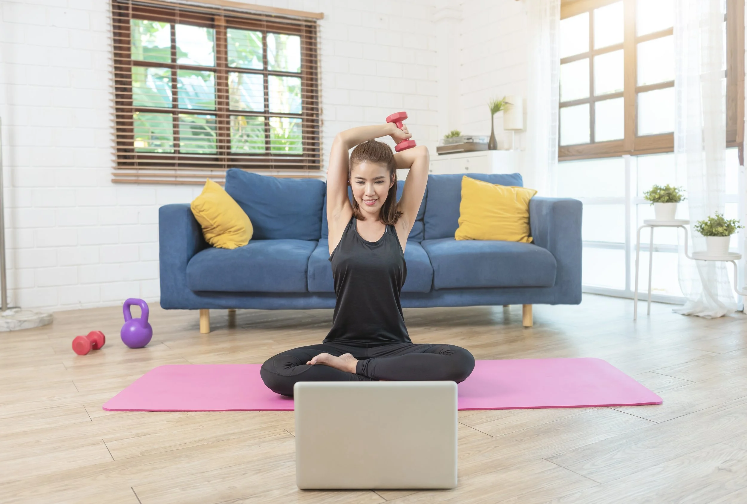 A woman exercises with dumbbells on a pink yoga mat in a bright living room, watching a workout video on her laptop.