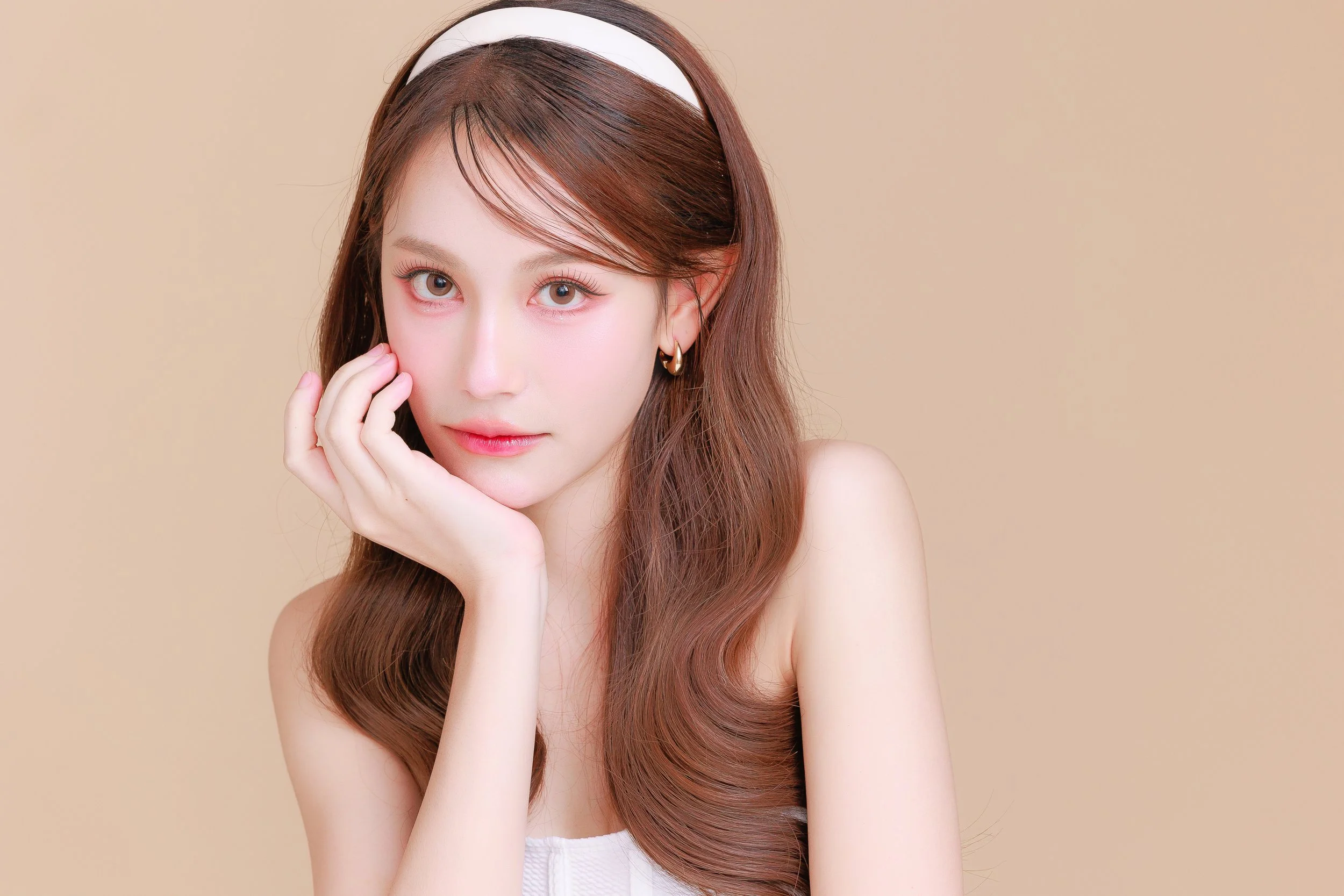 A young woman with long, wavy brown hair wearing a white headband, gazing directly at the camera with her hand resting on her face, against a neutral beige background.
