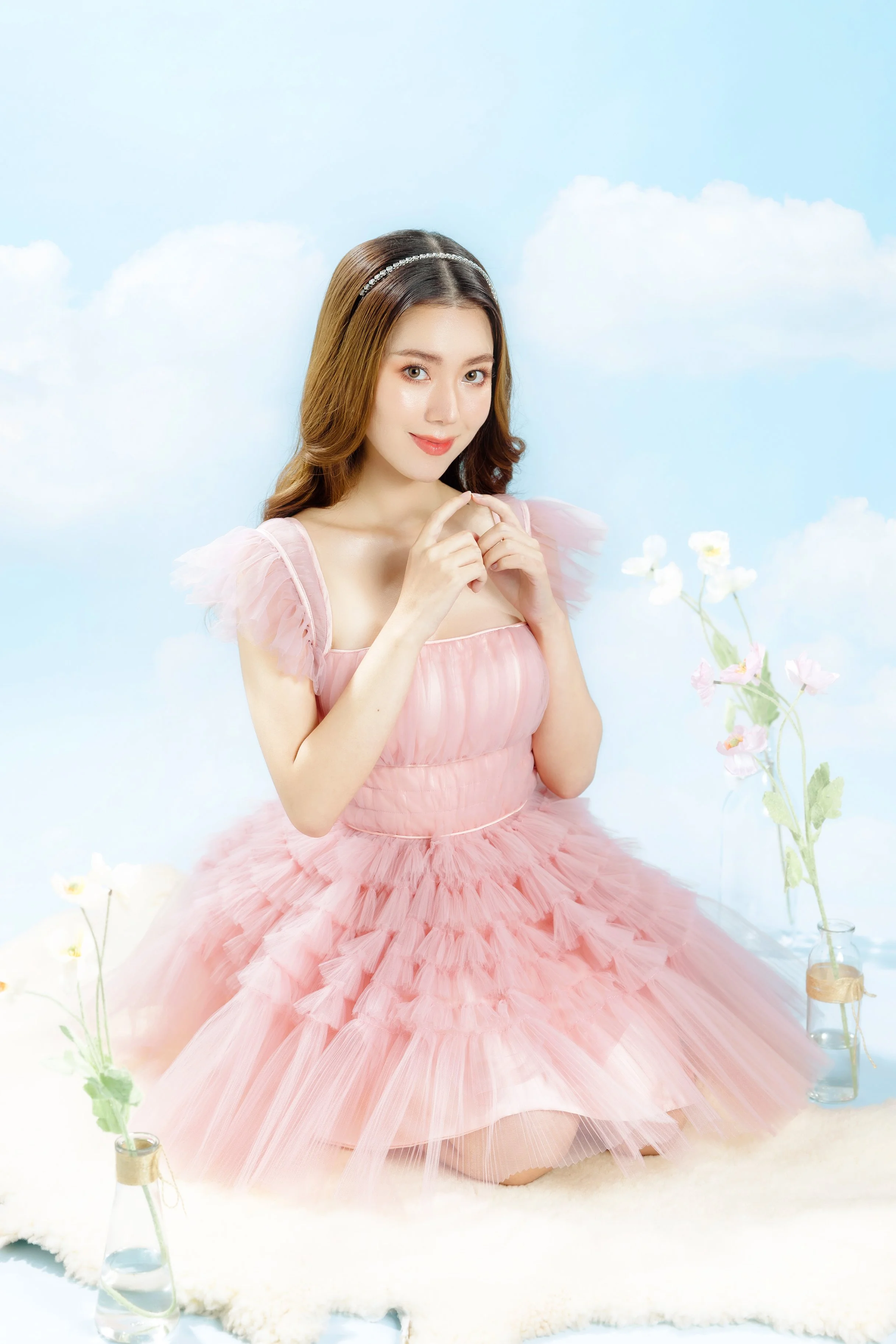 Young woman in a pink tulle dress with ruffled sleeves, sitting on a fluffy rug surrounded by glass bottles with flowers, against a light blue sky background with clouds.