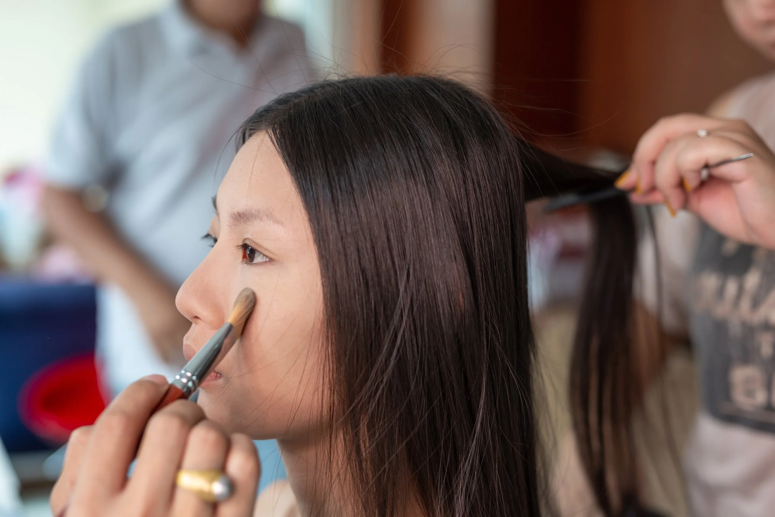 A woman getting her makeup done, with someone applying eyeshadow and another hairstylist working on her hair.