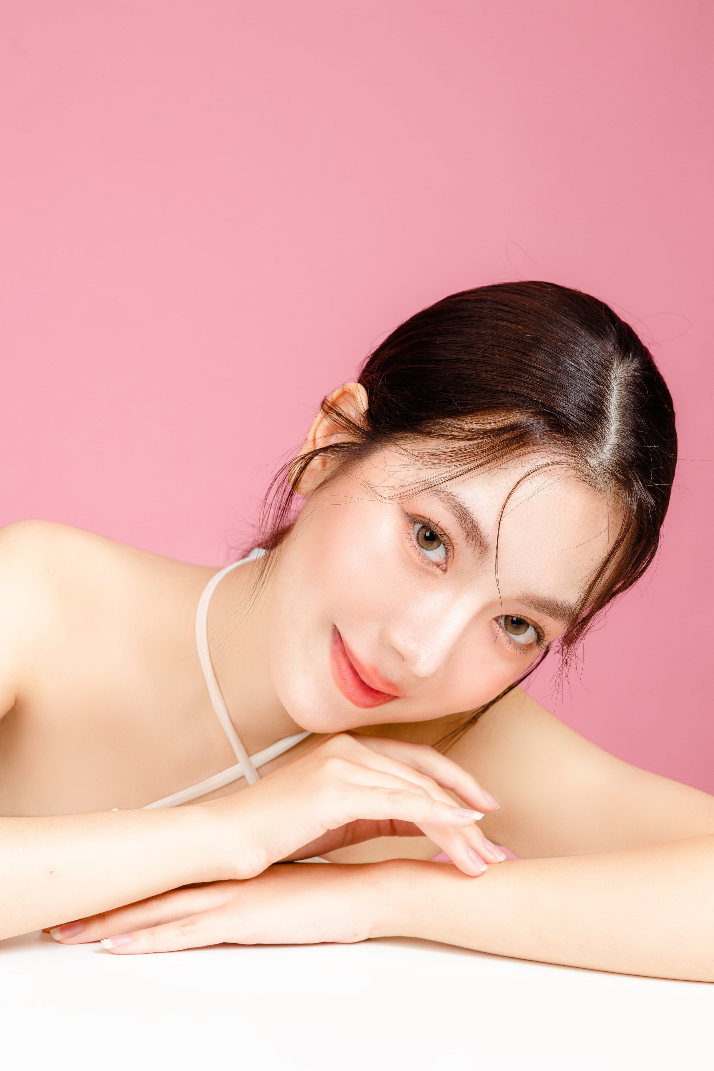 Close-up of a young woman with dark hair, light skin, and hazel eyes, smiling and resting her chin on her folded arms against a pink background.