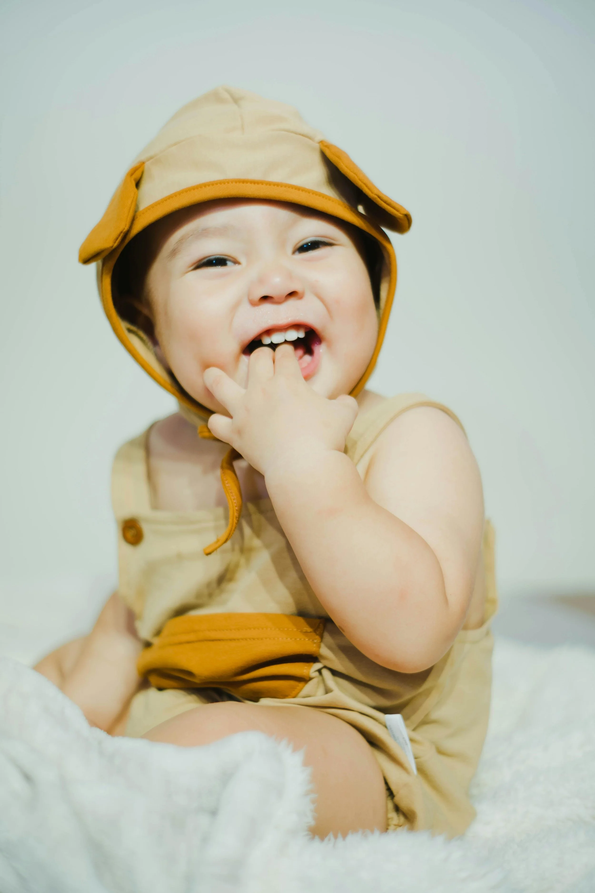 A young child smiling and looking at the camera, with fingers near their mouth, wearing a beige and orange sun hat and matching beige outfit, sitting on a white blanket against a plain background.