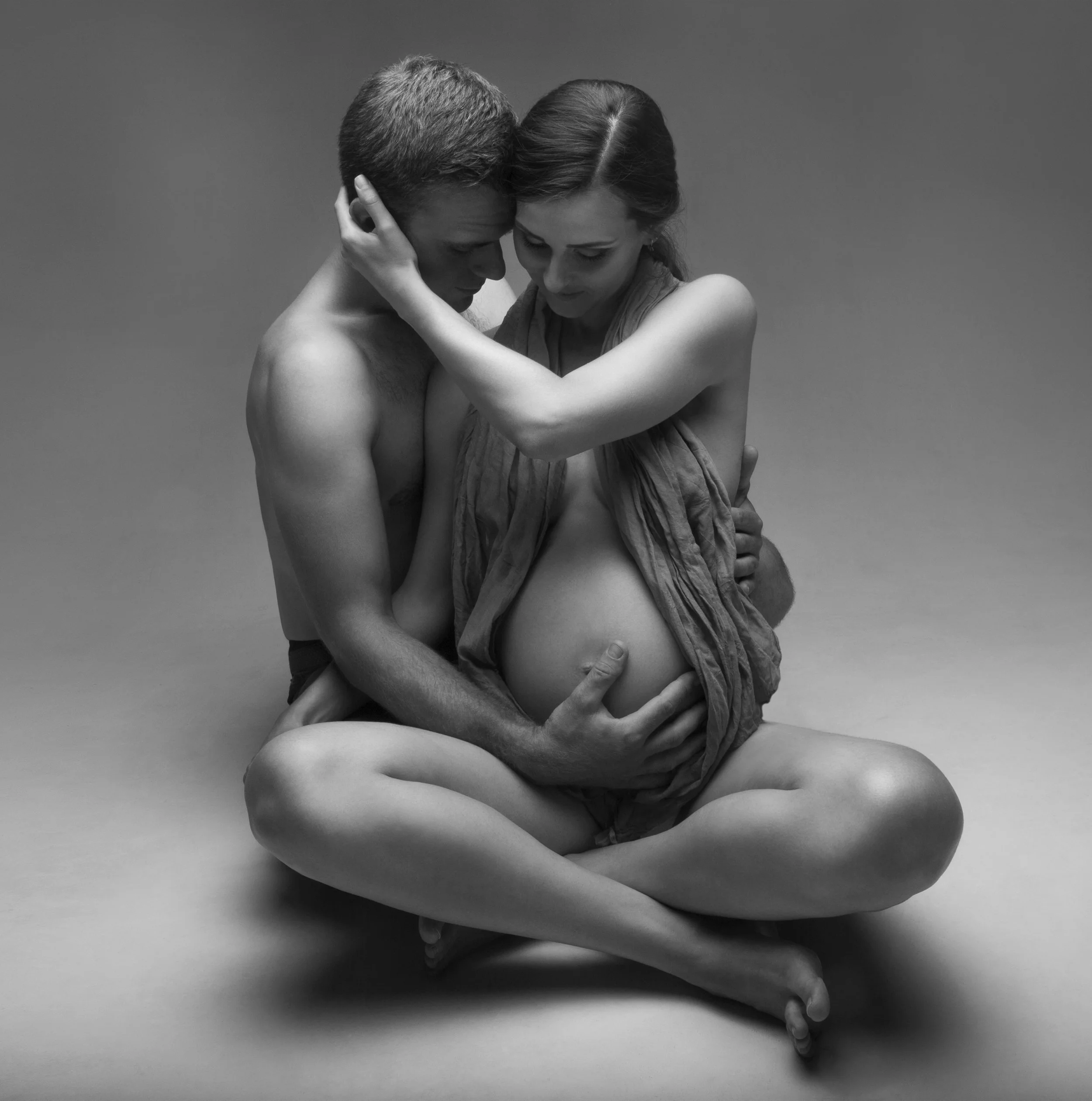 A black and white photo of a pregnant woman and a man sitting on the floor, embracing each other intimately.