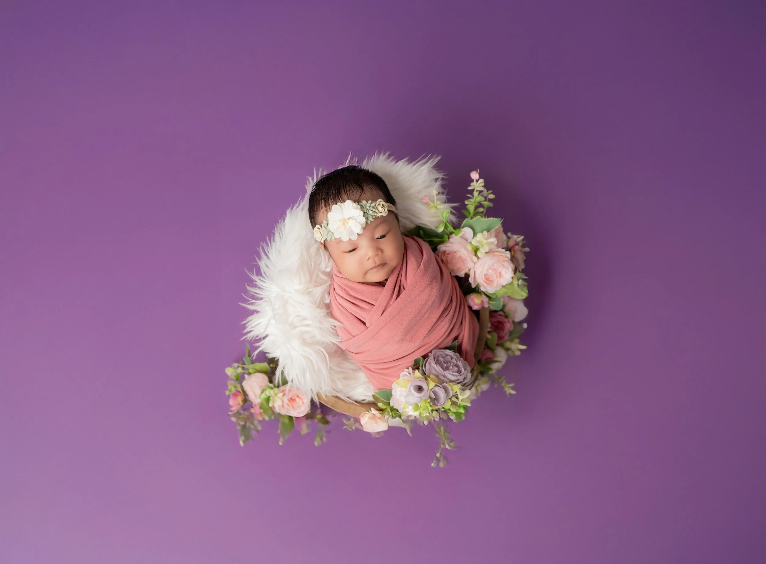 A newborn baby wrapped in pink fabric, wearing a flower headband, lying on a faux fur blanket surrounded by pink and purple flowers on a purple background.