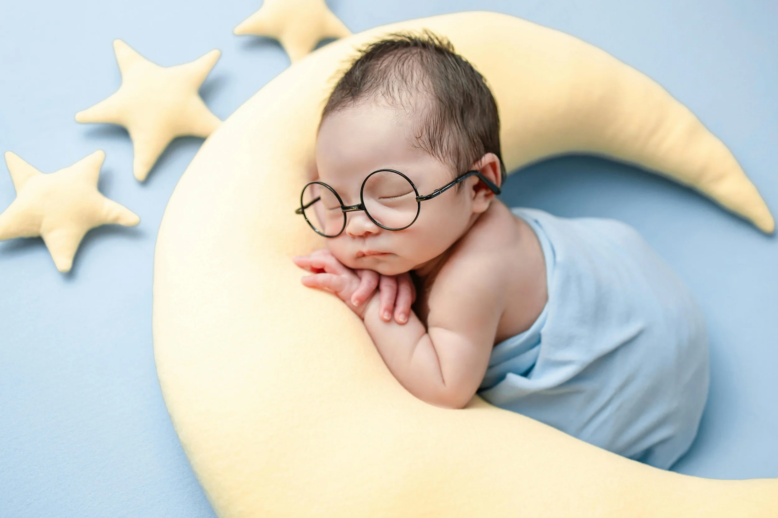 A sleeping baby with glasses, resting on a yellow crescent-shaped pillow, surrounded by plush star pillows on a blue background.