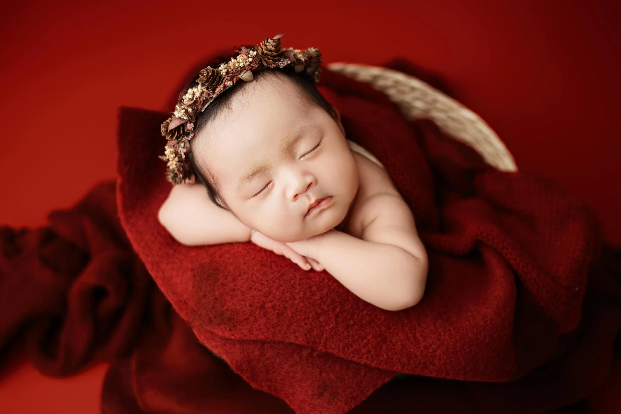 A sleeping newborn baby with a floral headband, lying on a red blanket, surrounded by a red backdrop.