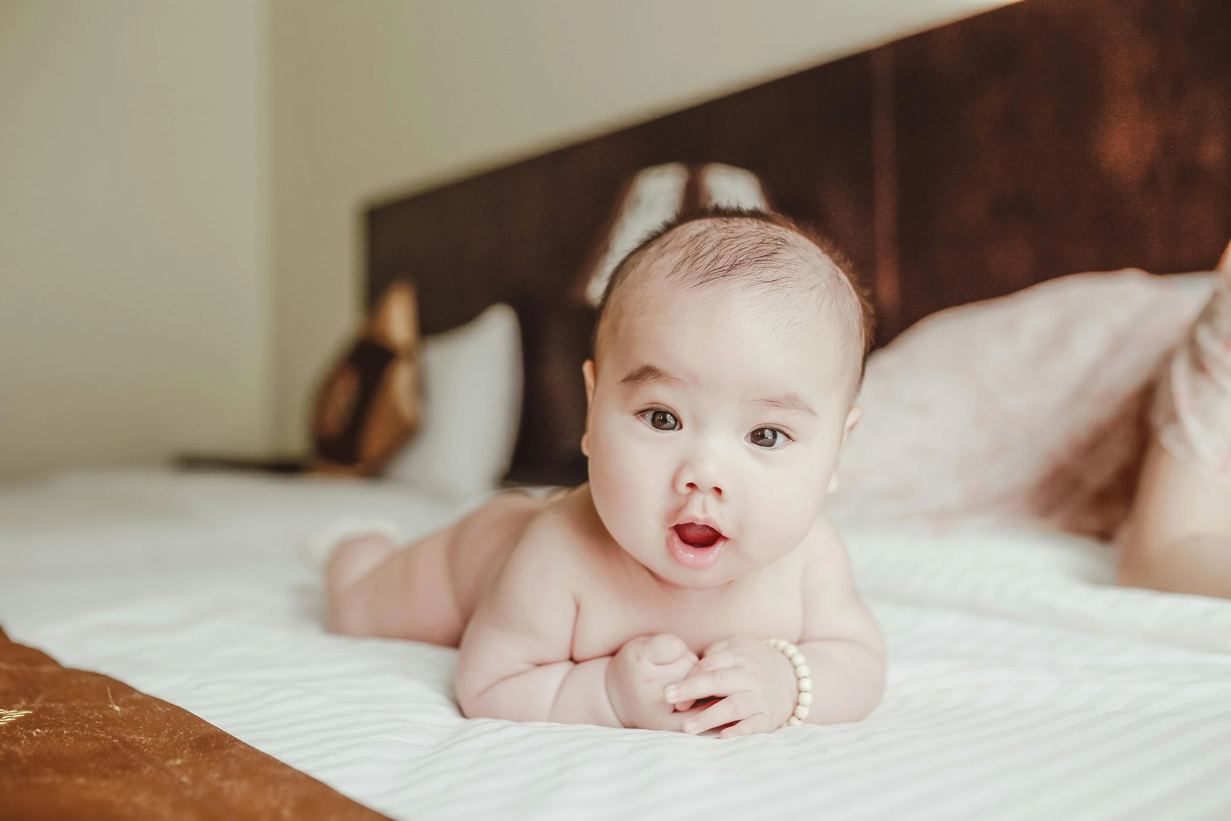 A baby with light skin and dark hair lying on stomach on a bed, looking at the camera with mouth slightly open.
