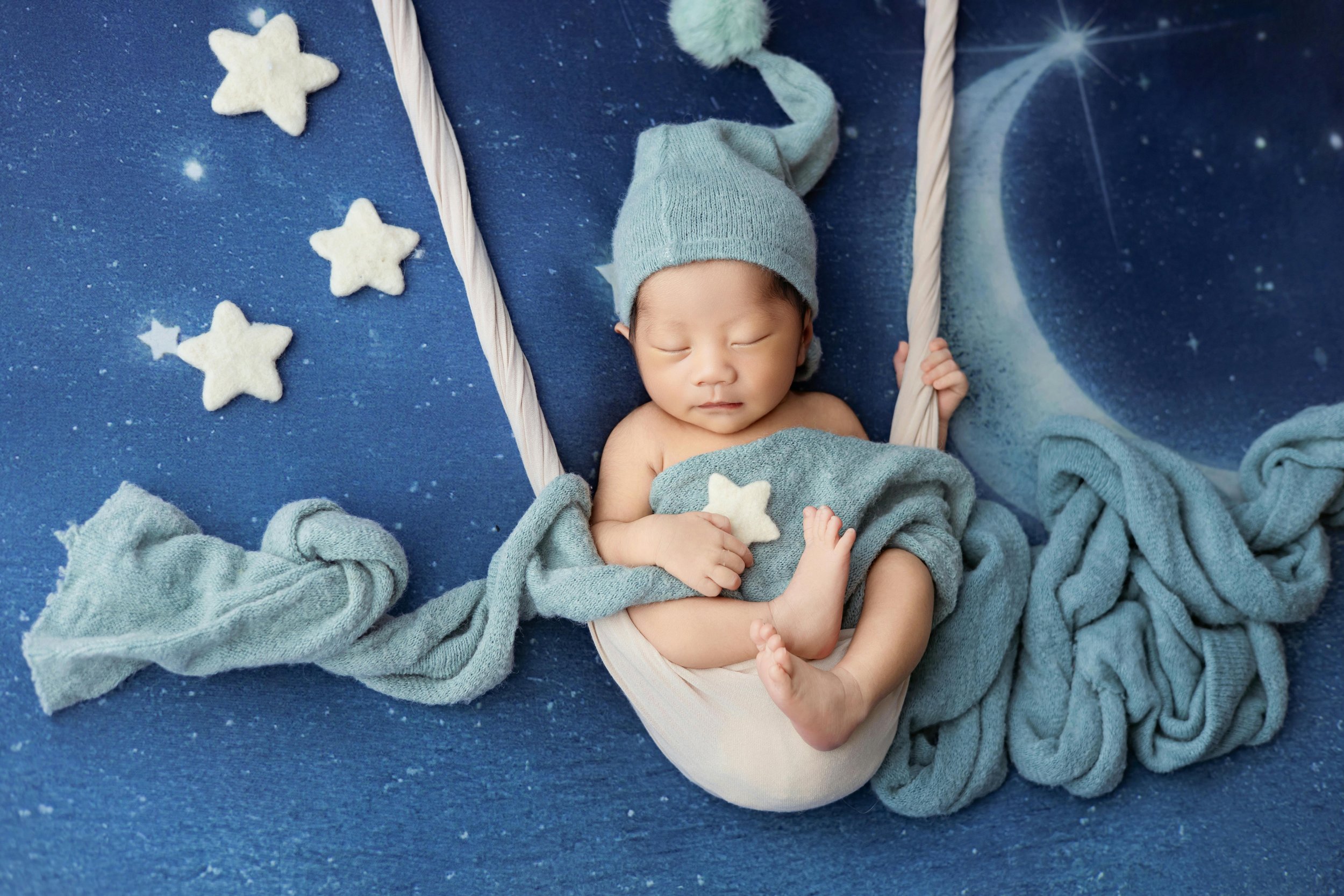 Sleeping baby wearing a gray hat, lying in a crib with blue star and moon-themed bedding, holding a star-shaped pillow, surrounded by soft blue blankets and star-shaped decorations.